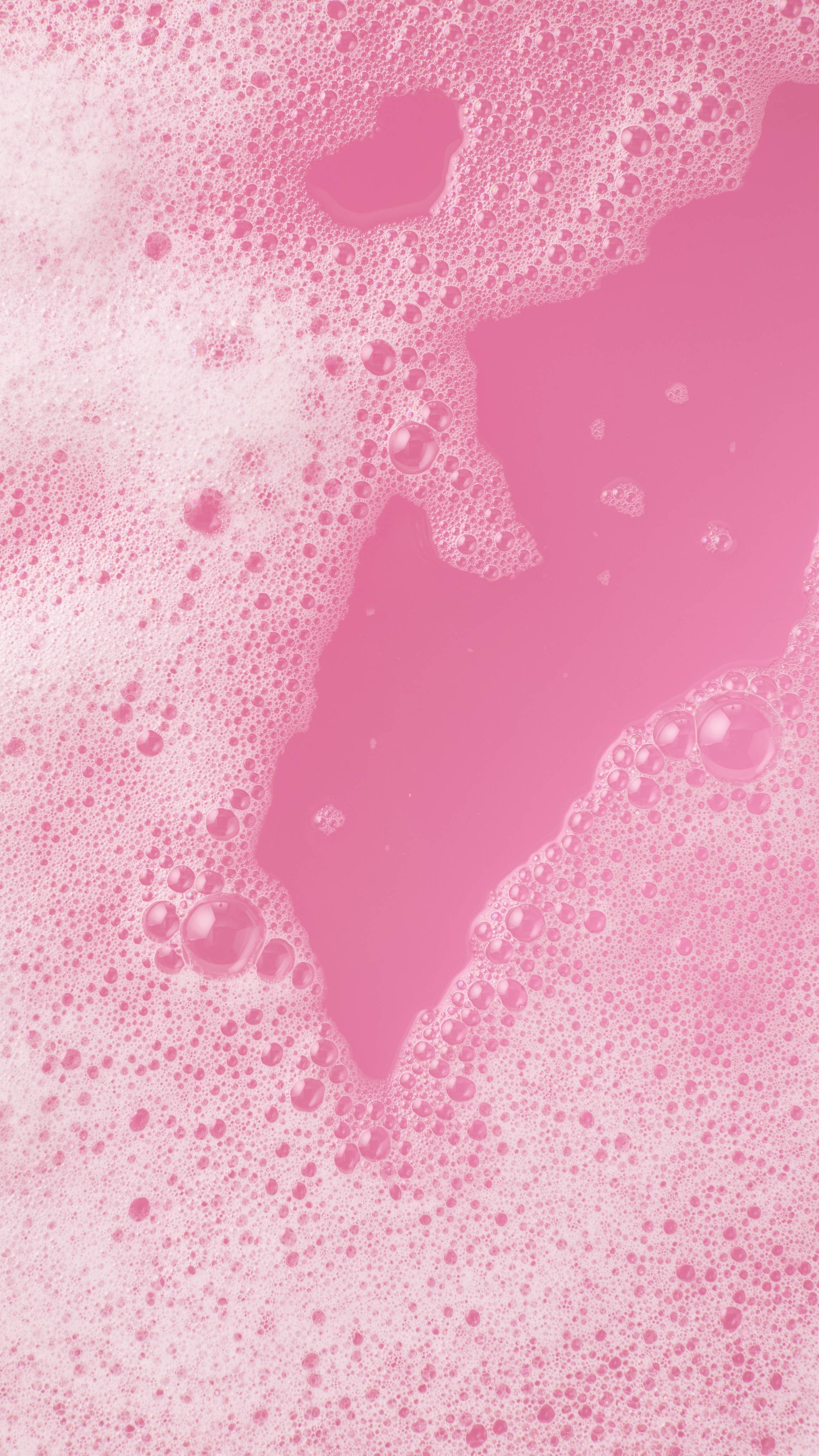 Top-down view of a bright pink bath covered in white, frothy bubbles. The vibrant pink water is visible through a large clearing in the center of the foam.