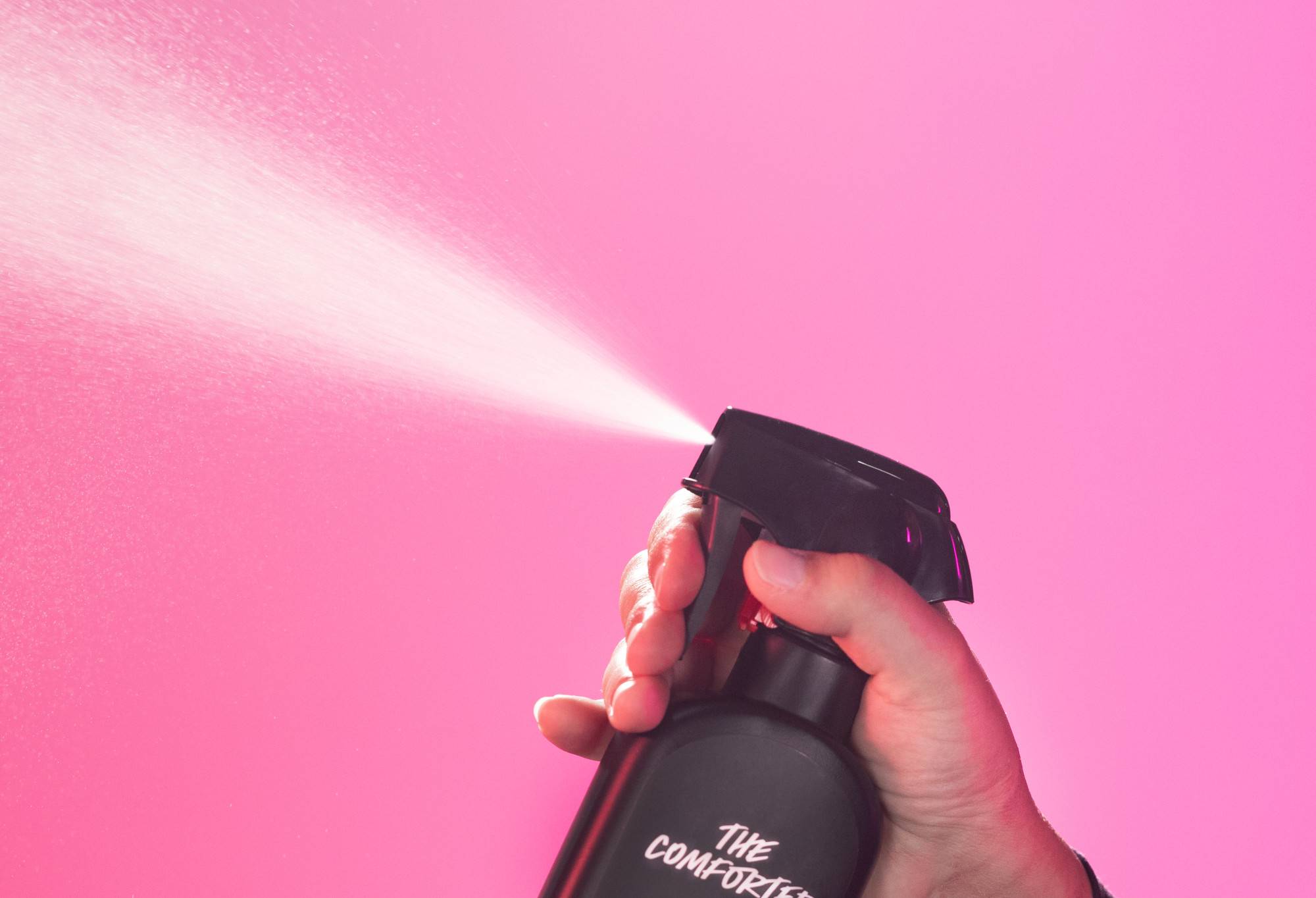 The Comforter body spray is sprayed up into the air, in front of a vibrant, candy pink background.