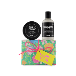 Comfy Cosy. A short rectangular gift box wrapped in pastel-coloured paper designed with flowers in a hand-drawn style, tied with a pink string and yellow tag. Two themed products sit on top of the box.