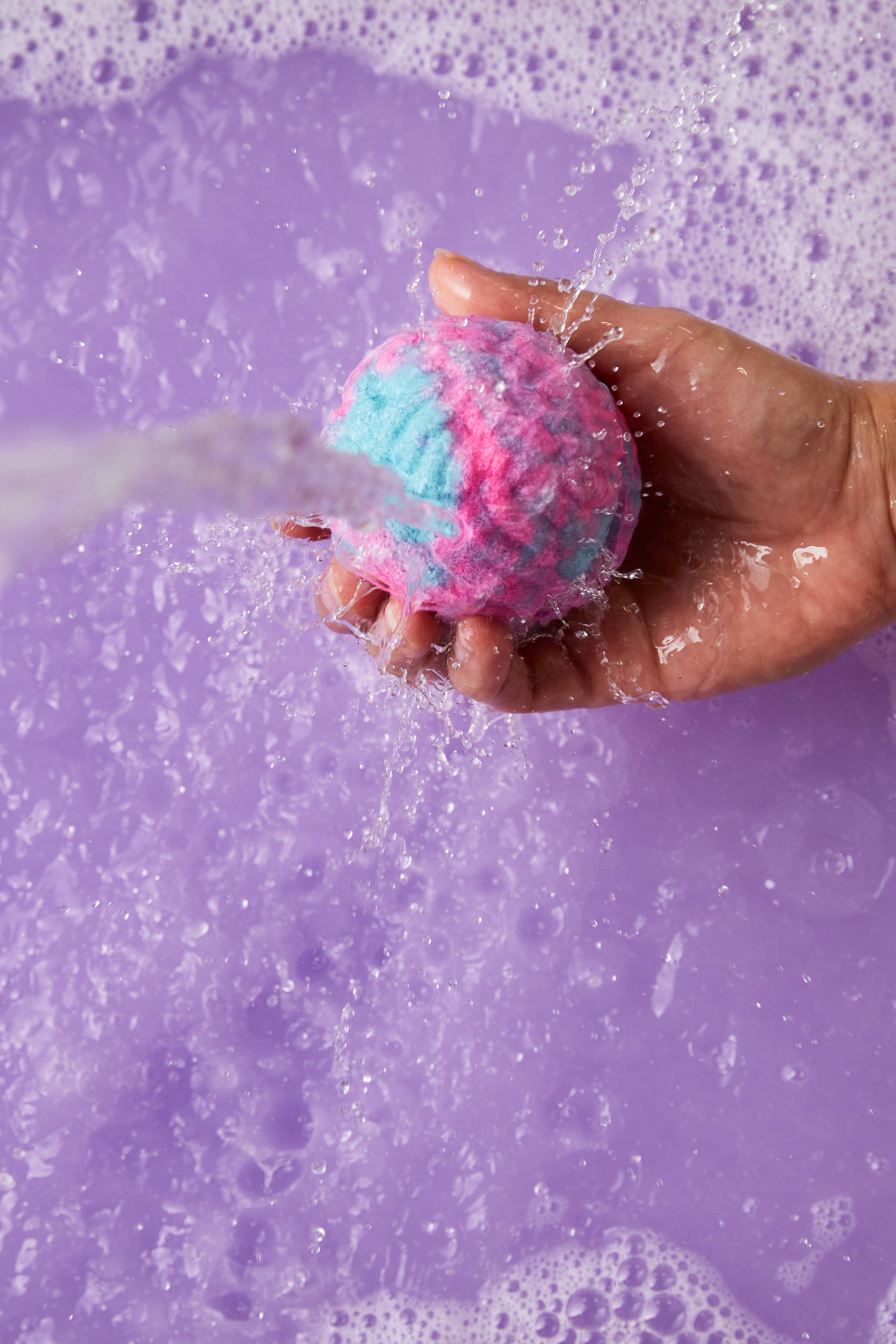 The image shows a light-toned hand holding a marbled pink, purple, and light blue Concha Cubbleroon bubble bar under a strong stream of water. Splashes and purple foam erupt into the light purple bath.