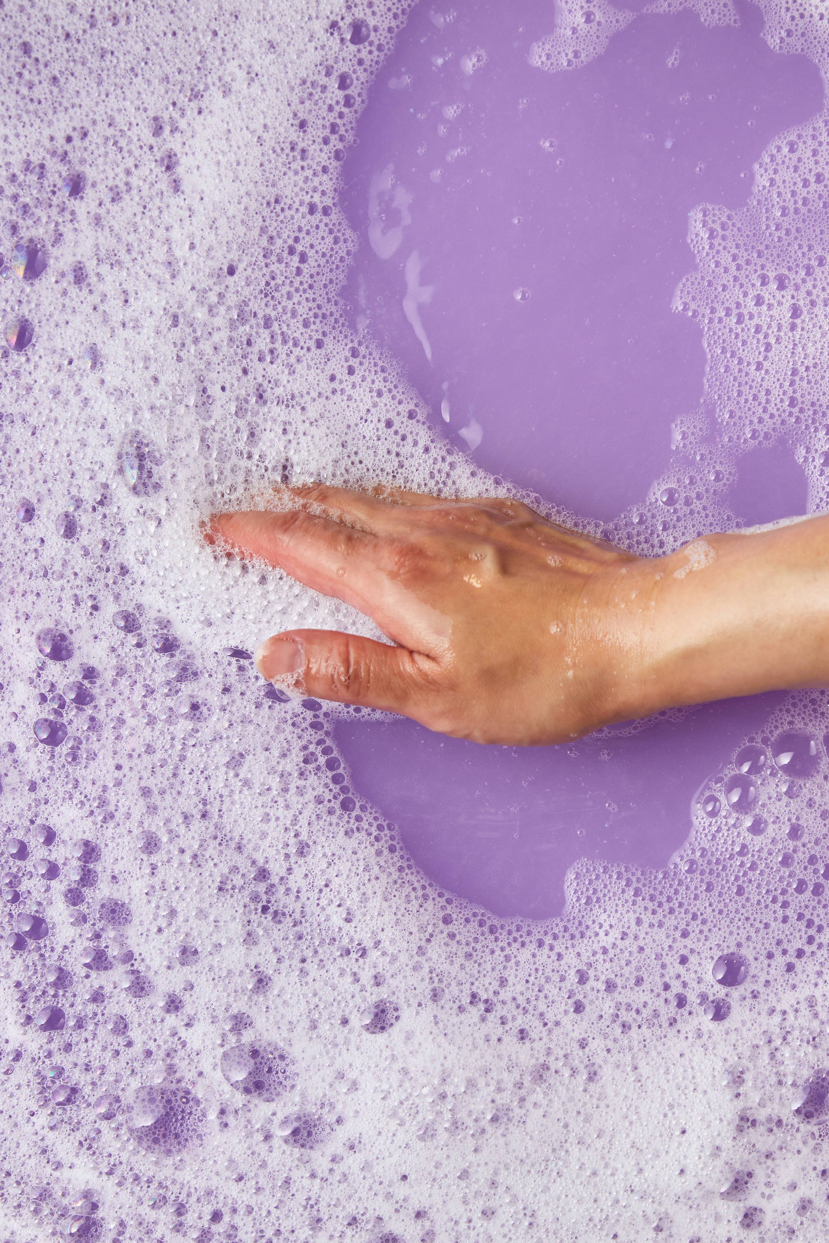 The image shows a light-toned hand gently touching the surface of thick white foam, parted to reveal a pool of smooth purple bath water. Small bubbles float in both the foam and the water, made by the Concha Cubbleroon bubble bar.