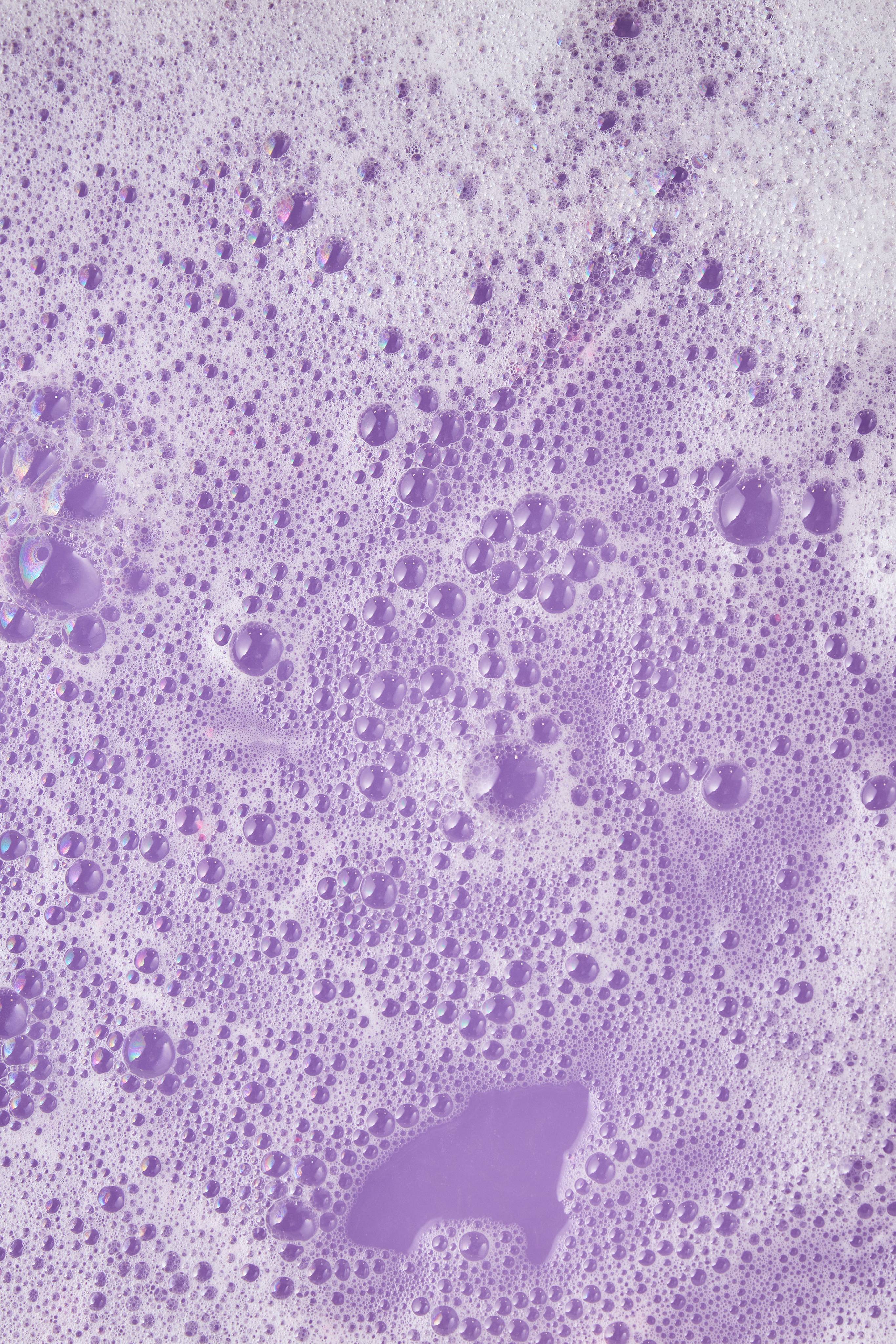 The image shows a vast surface of lilac foam with numerous bubbles of all sizes. A smooth puddle of deep purple liquid forms an irregular shape in the centre, creating an abstract, bubbly landscape.