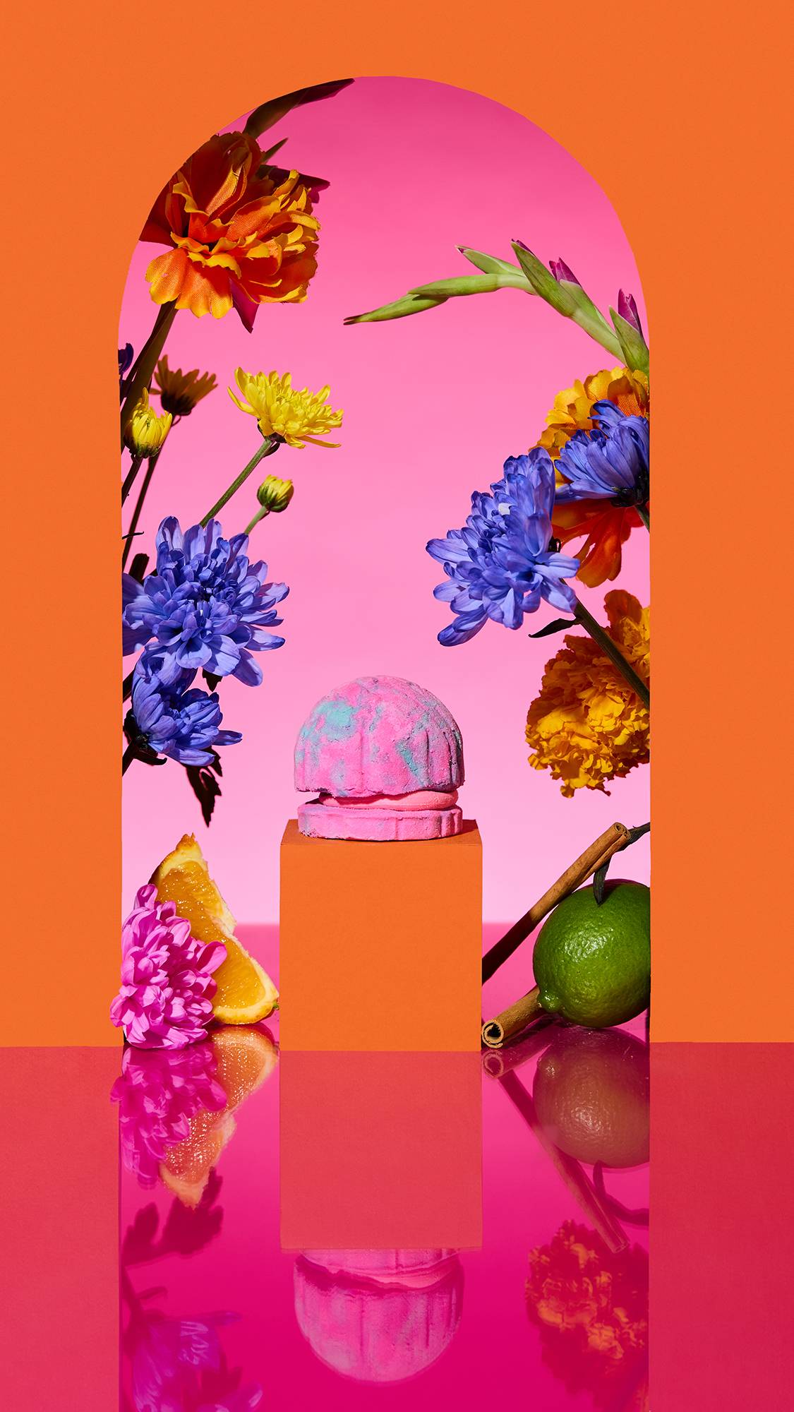 The image shows a marbled pink and blue 'Concha Cubbleroon' bubble bar on a plinth. A colourful arch frames the scene, with sliced citrus, flowers, and a cinnamon stick surrounding the product on a reflective pink surface.