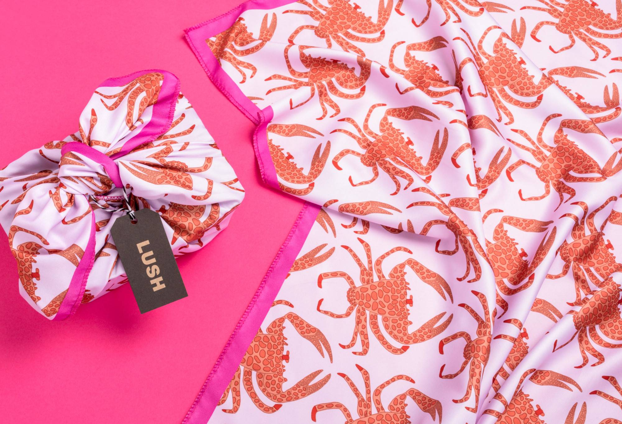 The knot wrap is wrapped around a rectangular box with a tag, as well as laid out flat, on a bright pink background.