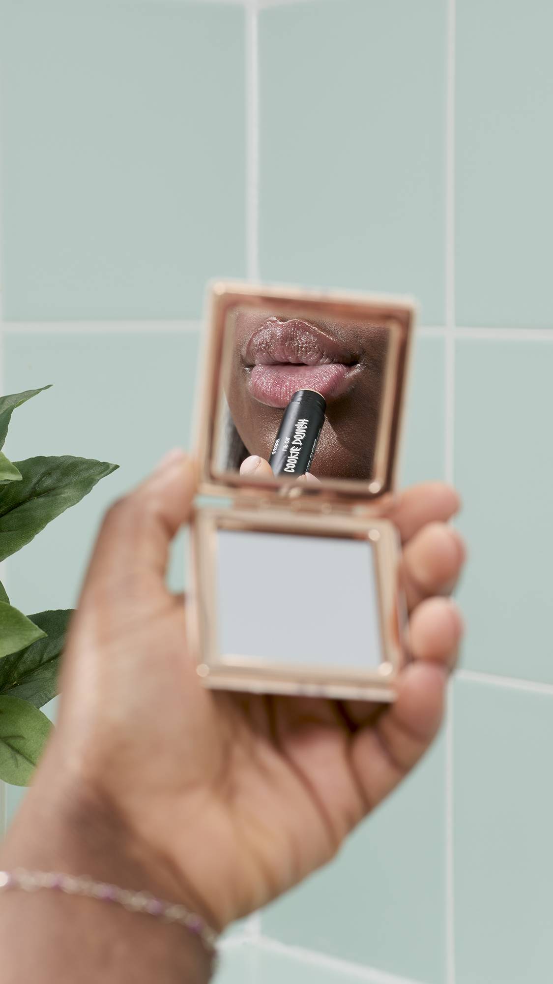A close-up shot shows a hand holding a gold compact mirror. The reflection shows a model's lips while applying Lush 'Cookie Dough' lip balm. The background features light blue bathroom tiles and a glimpse of green foliage.