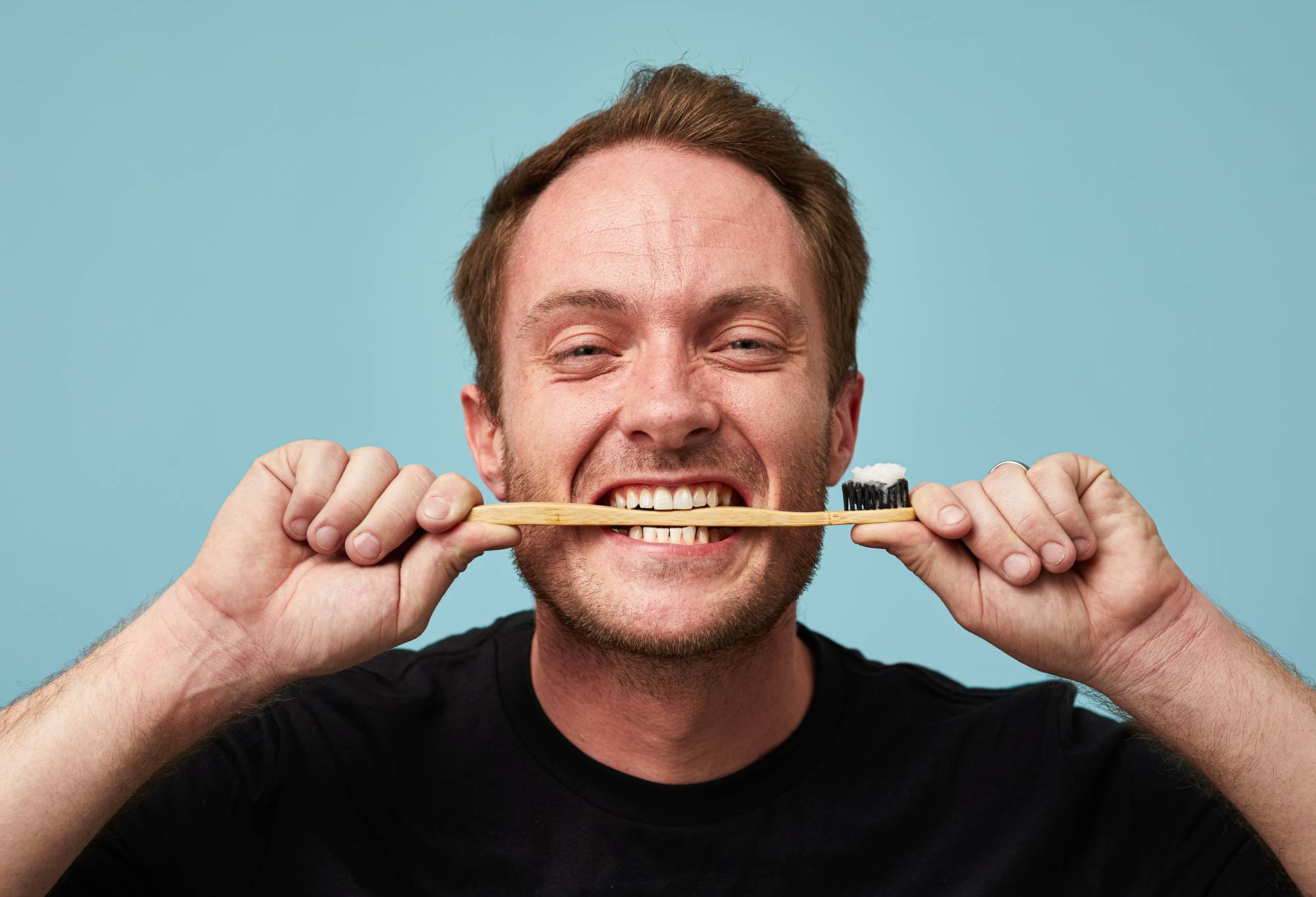 A model holds a toothbrush horizontally between their teeth, with white toothpaste jelly on the bristles. 