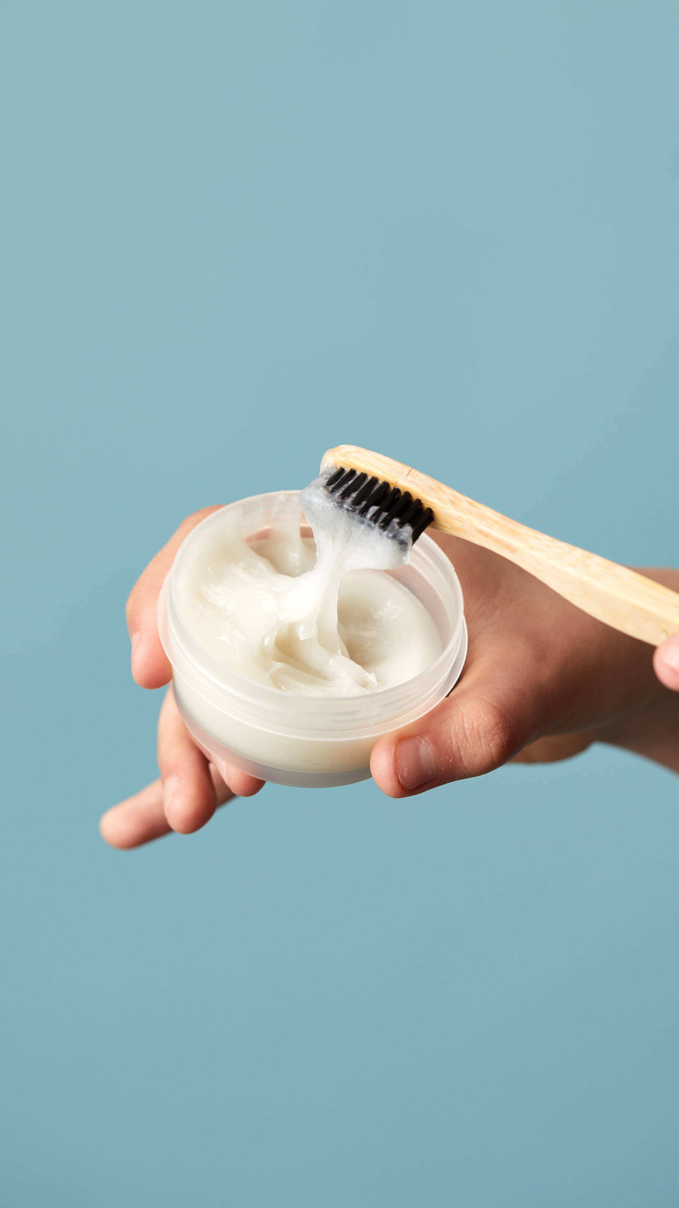 The model's hands hold the pot of toothpaste while dipping a bamboo toothbrush into the paste on a pale blue background.
