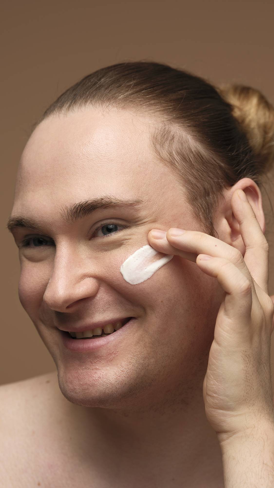 A close-up image of the model's smiling face as they start to apply the Cosmetic Lad self-preserving moisturiser to one cheek. They are on a warm, earthy-brown background and their hair is tied back.
