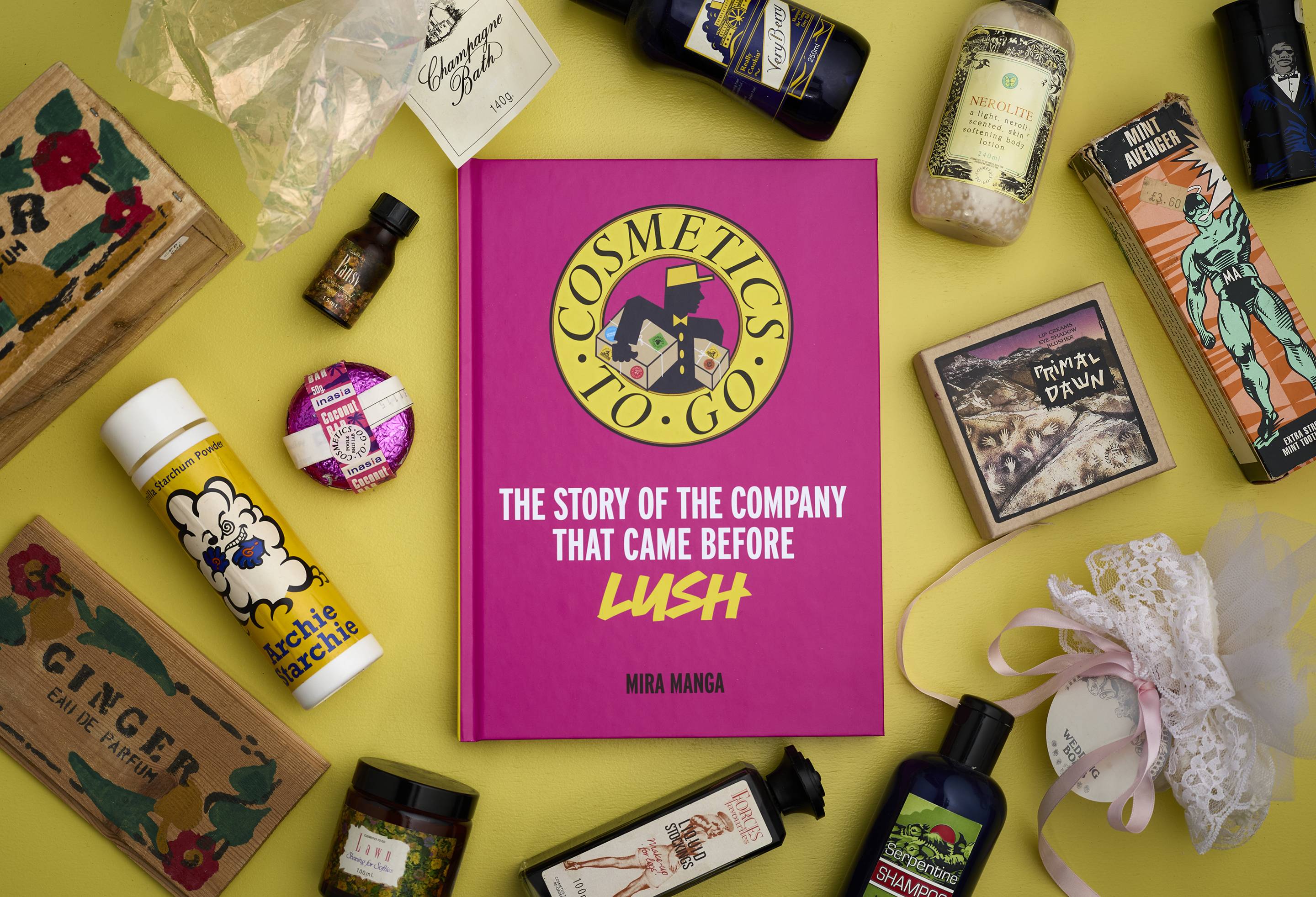 An aerial shot of the Cosmetics To Go book. The book is pink with a yellow circle logo and rests on a yellow background, surrounded by various vintage Cosmetics To Go products, including perfumes, bath bombs, and bath oils.