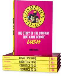  A stack of four hardcover books sit with an open book on top. Its front cover is bright pink with a yellow circle with the words "COSMETICS TO GO" and a logo inside. The words "THE STORY OF THE COMPANY THAT CAME BEFORE LUSH" are underneath.