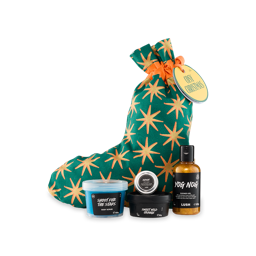 Cosy Christmas. A cotton fabric Christmas stocking-shaped gift bag. The gift bag is green with golden stars and a yellow gift tag. Four included Lush products sit in front of the bag. 