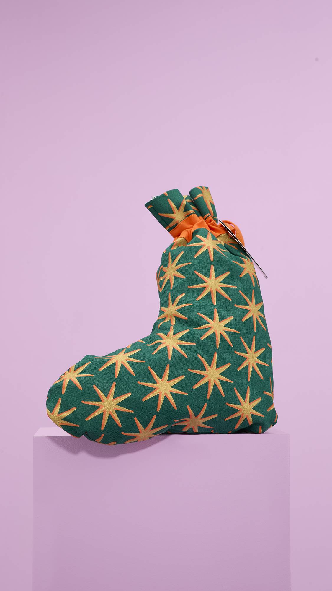 The image shows the green and starry, Christmas stocking-shaped gift bag sitting on a pale-pink platform against a matching pink background. 