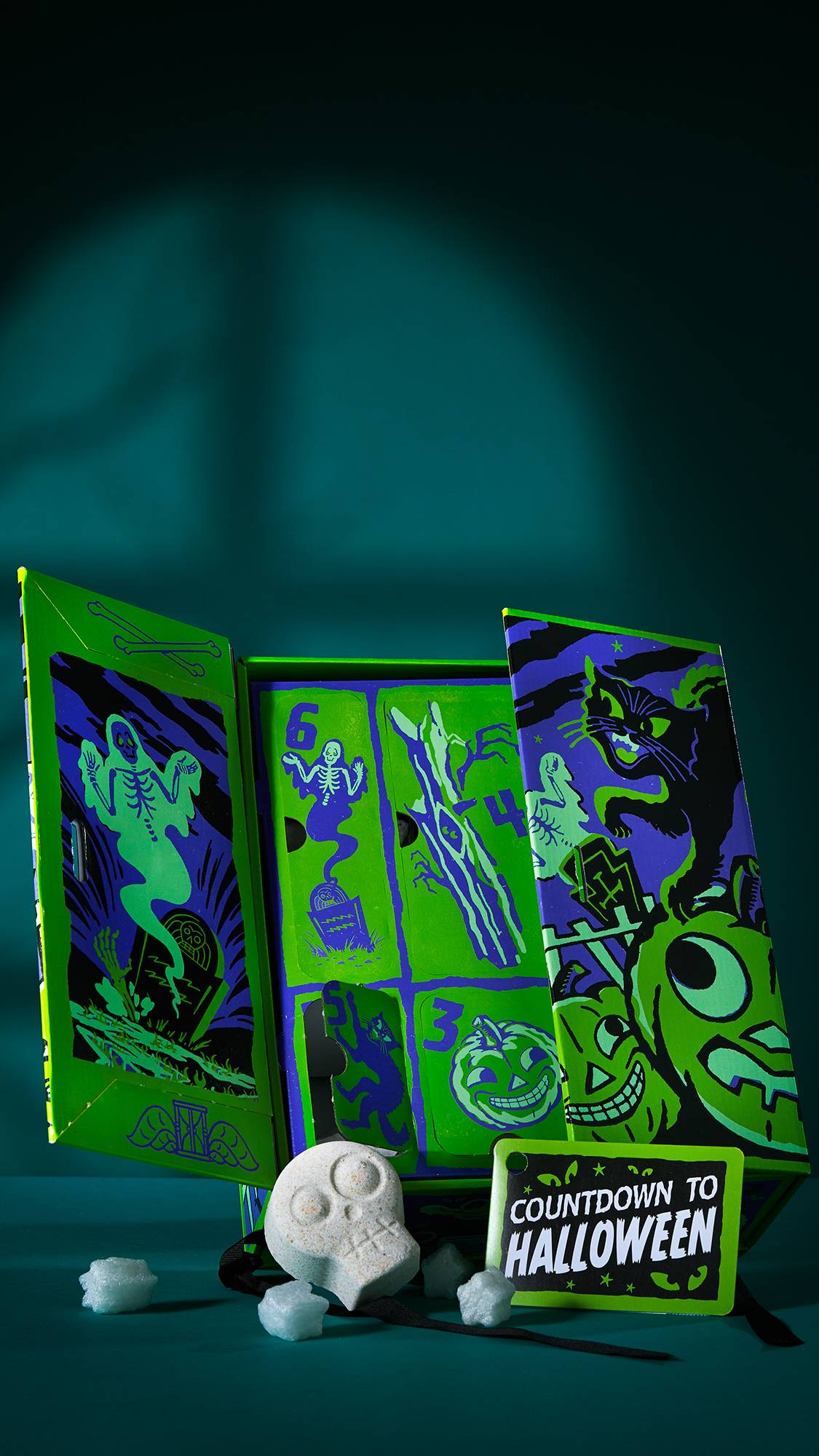 The image shows an open 'COUNTDOWN TO HALLOWEEN' gift box. The colourful box reveals numbered compartments inside. A white skull bath bomb and foam pieces sit in front of one of the open doors, against a dark teal background with shadows.