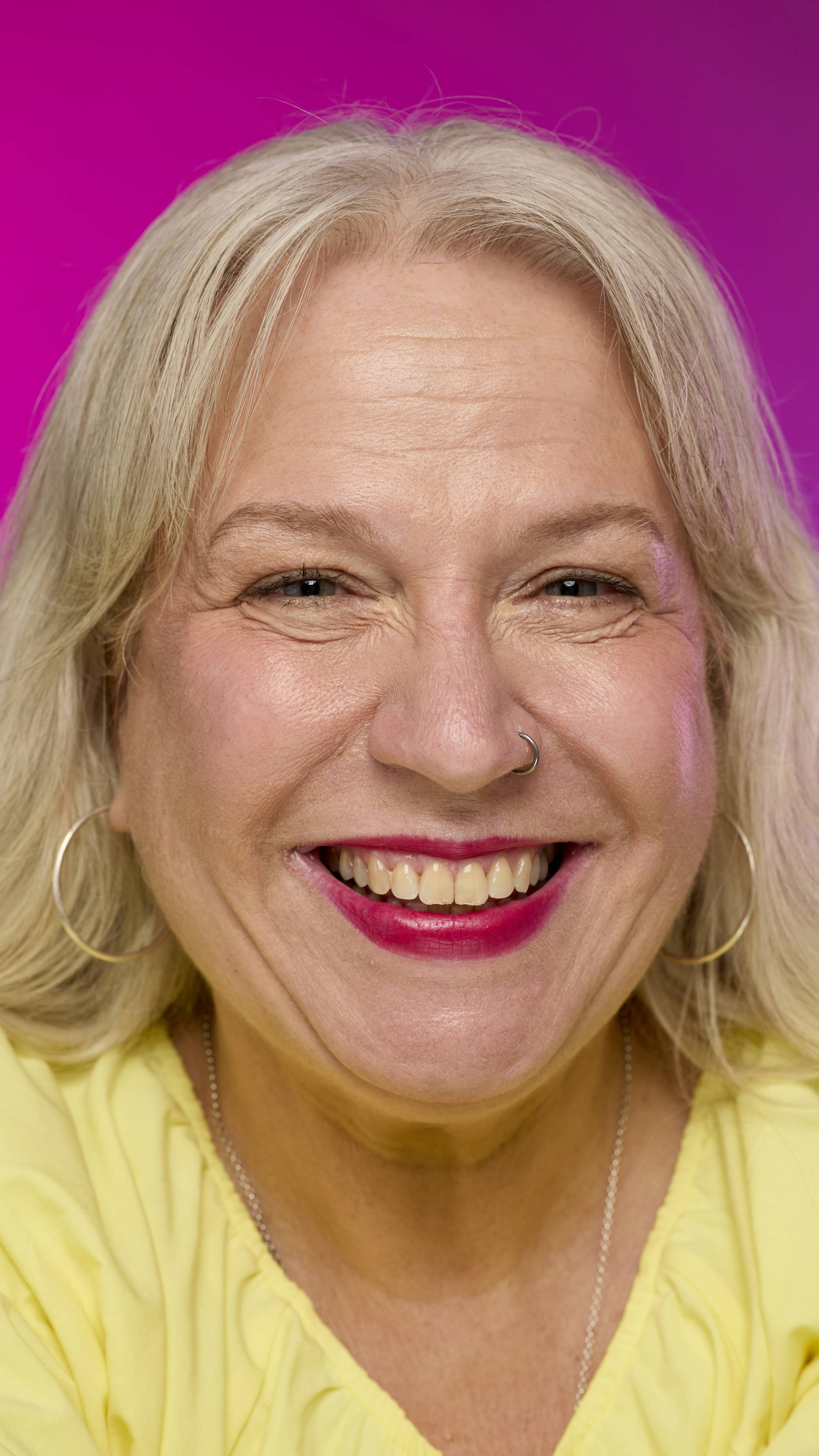 A smiling model with fair skin and light hair is shown from the neck up on a vibrant pink background. They smile, showing off the rich, crimson red Crackle lipstick with a subtle shimmer. 