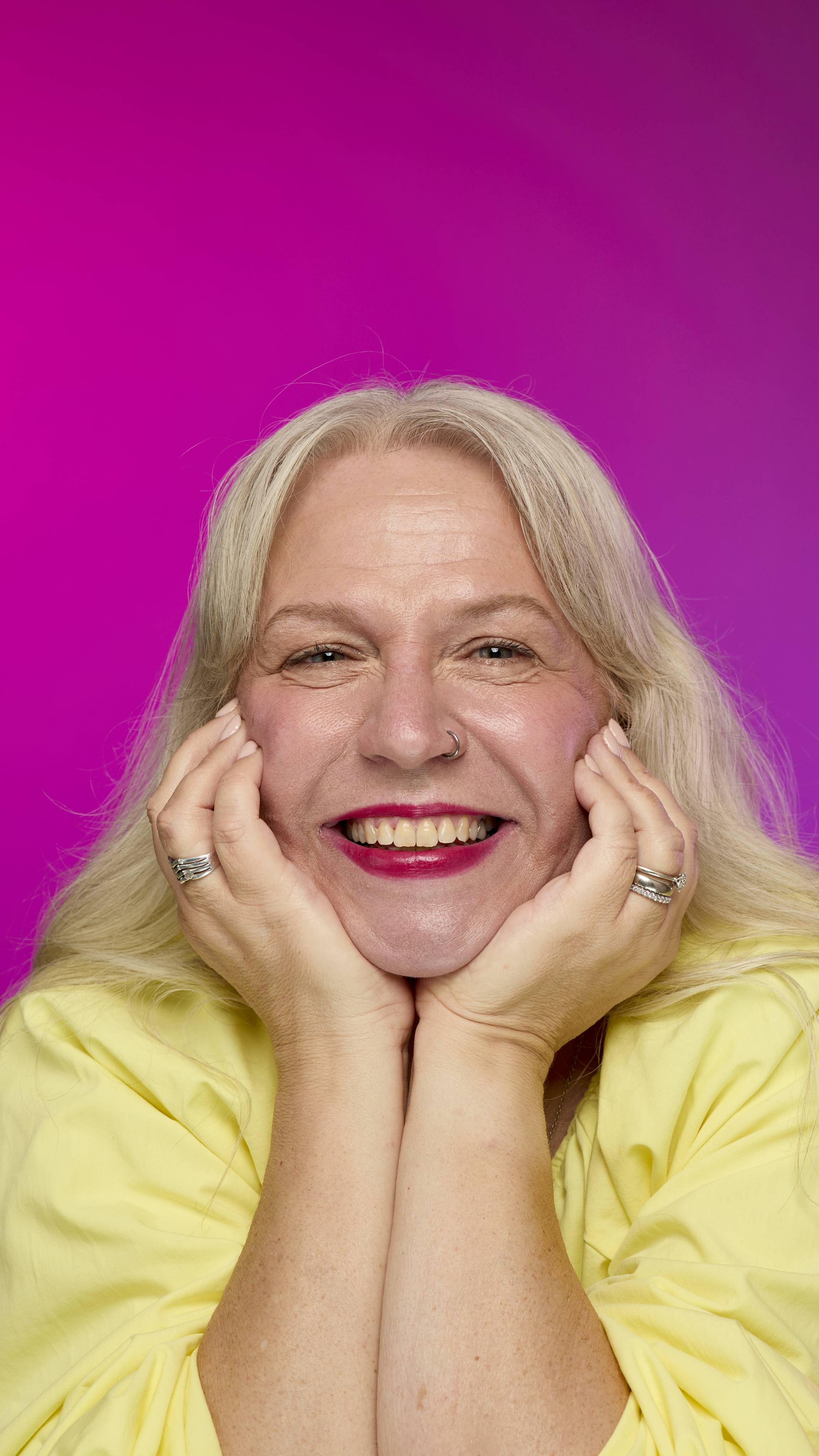 A smiling model with fair skin and light hair is shown from the neck up, with their chin resting on their hands, on a vibrant pink background. They smile, showing off the rich, crimson red Crackle lipstick with a subtle shimmer. 
