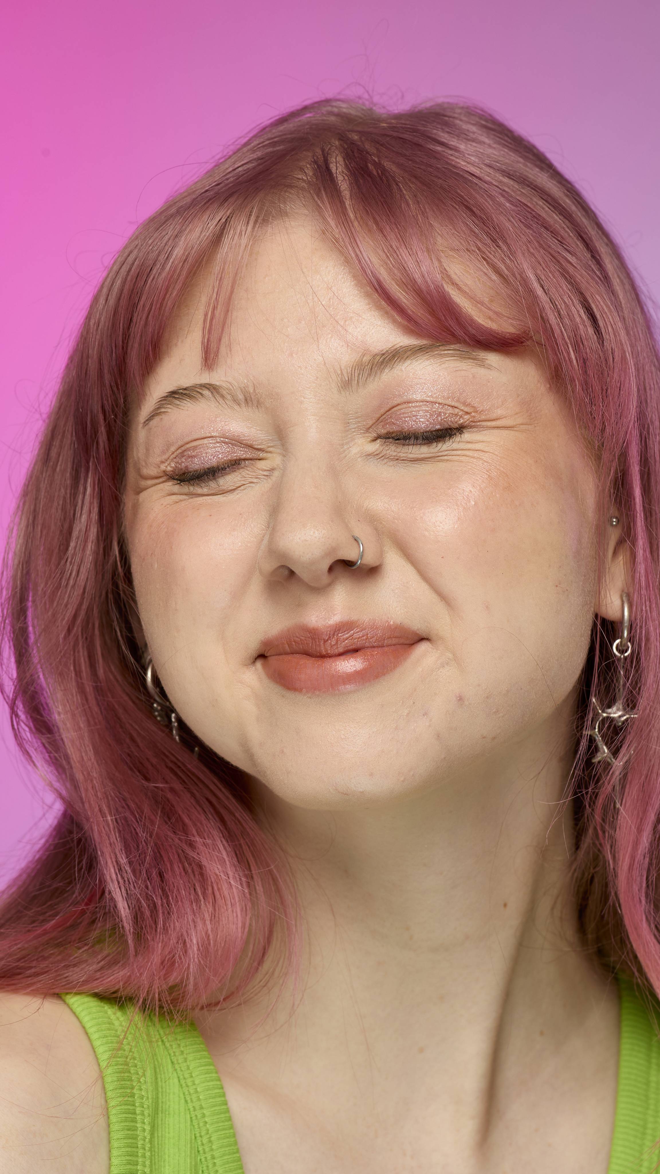 The image shows a fair-skinned model on a pink and blue gradient background, smiling with their eyes closed. They have pink hair and subtle shimmering lips from using the Crackled Copper lip cream. 