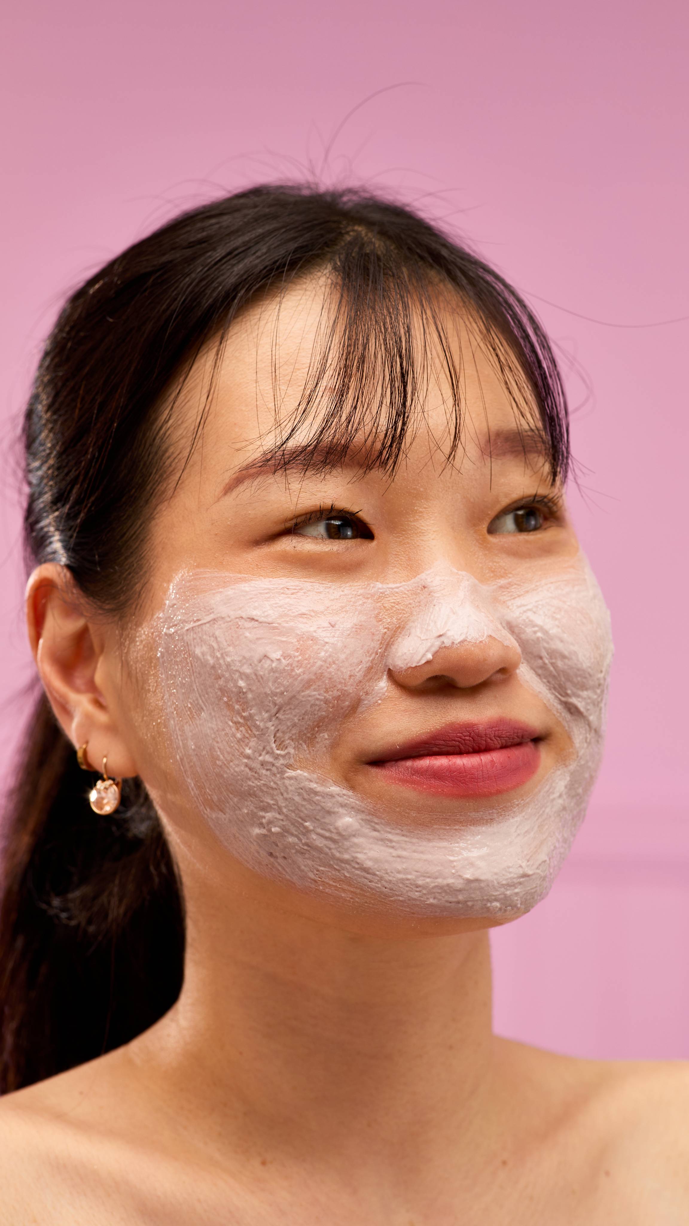 A person smiles with a light-coloured LUSH Cranberry jelly mask applied to their chin, cheeks, and nose. The mask has a textured, airy appearance, and the background is a soft pink.