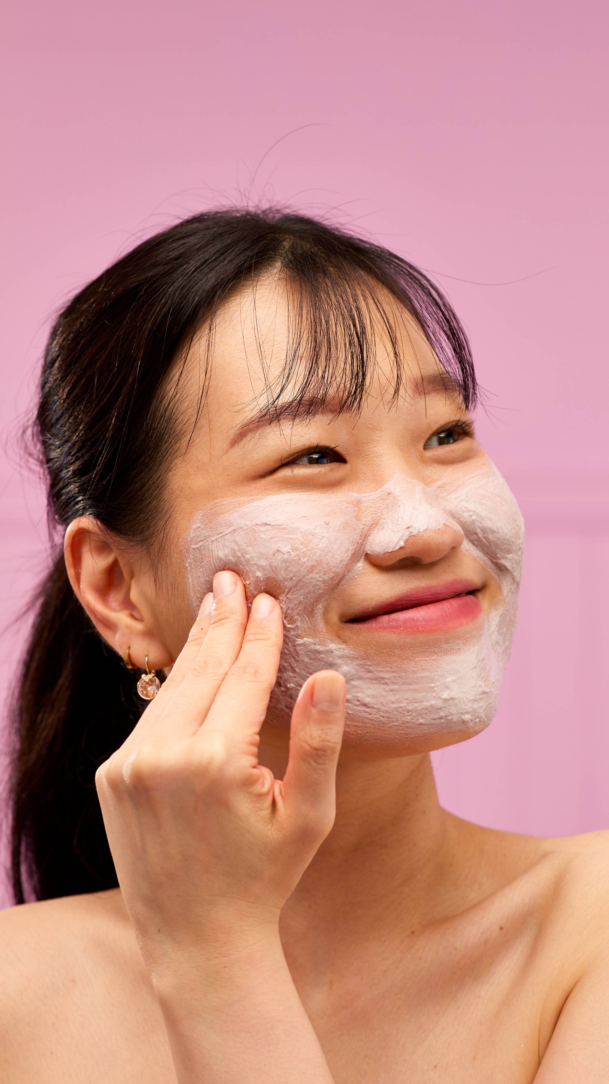 A person smiles as they apply a LUSH Cranberry jelly mask to their face. The textured, light-coloured mask covers their cheeks and chin, and the background is a soft pink.