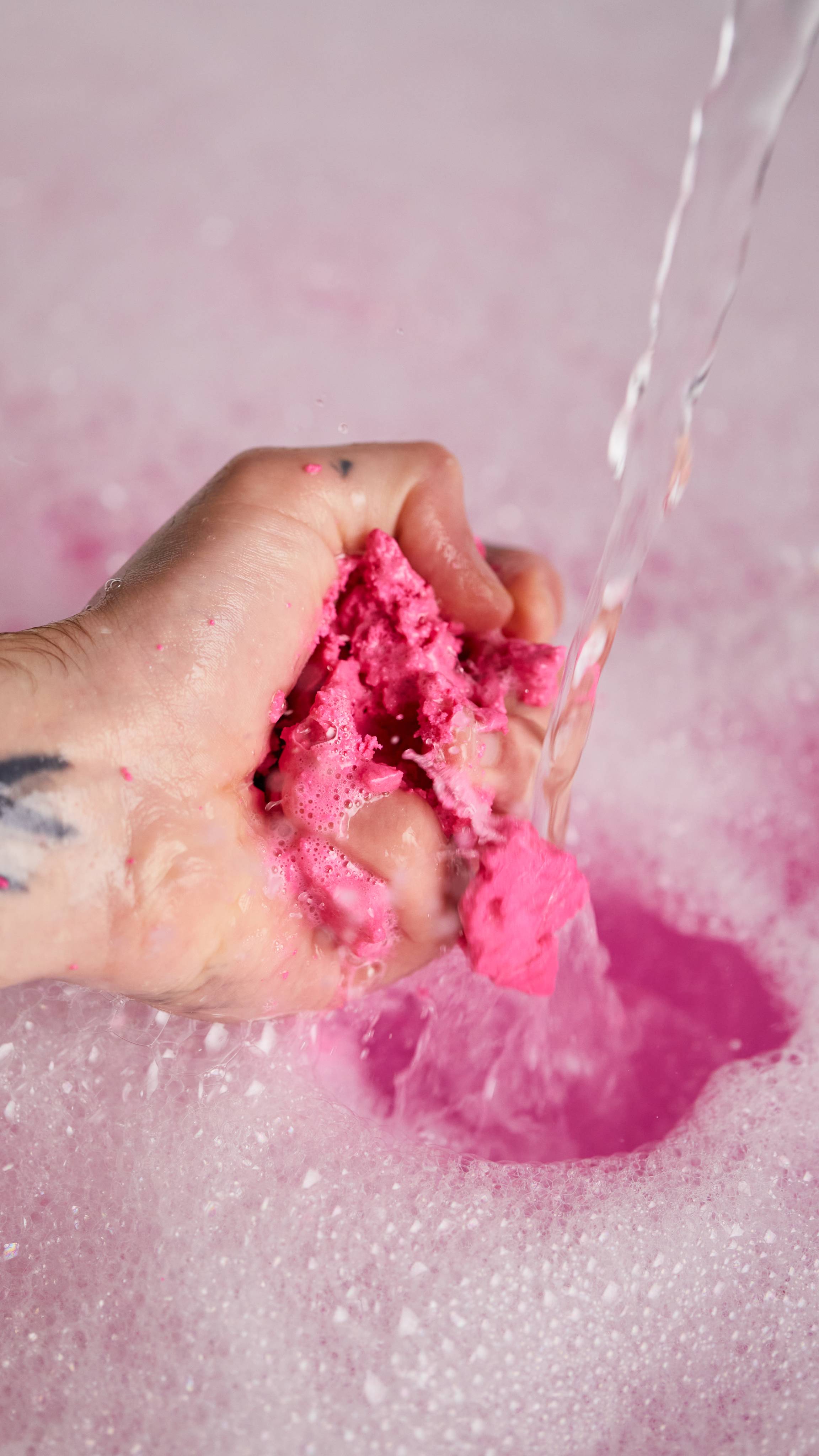 Image shows the model gently crumbling the pink bubble bar under running water in front of bubbly, pink bath water.