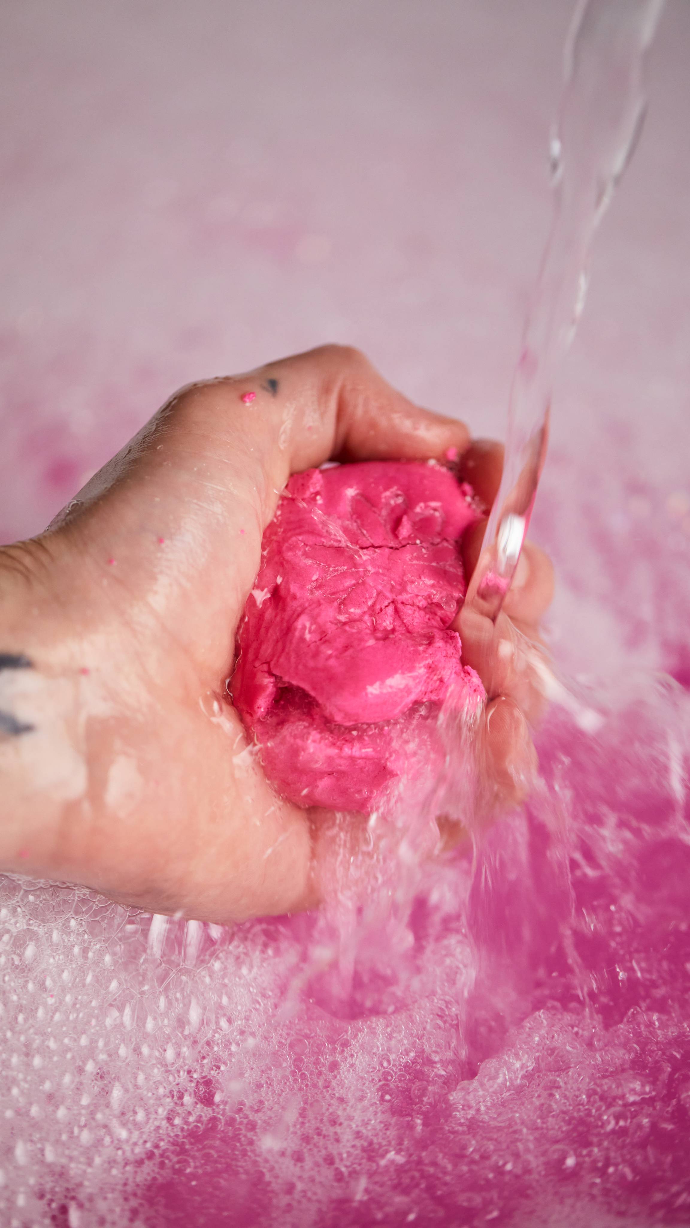 Image shows the model gently crumbling the pink bubble bar under running water in front of bubbly, pink bath water.