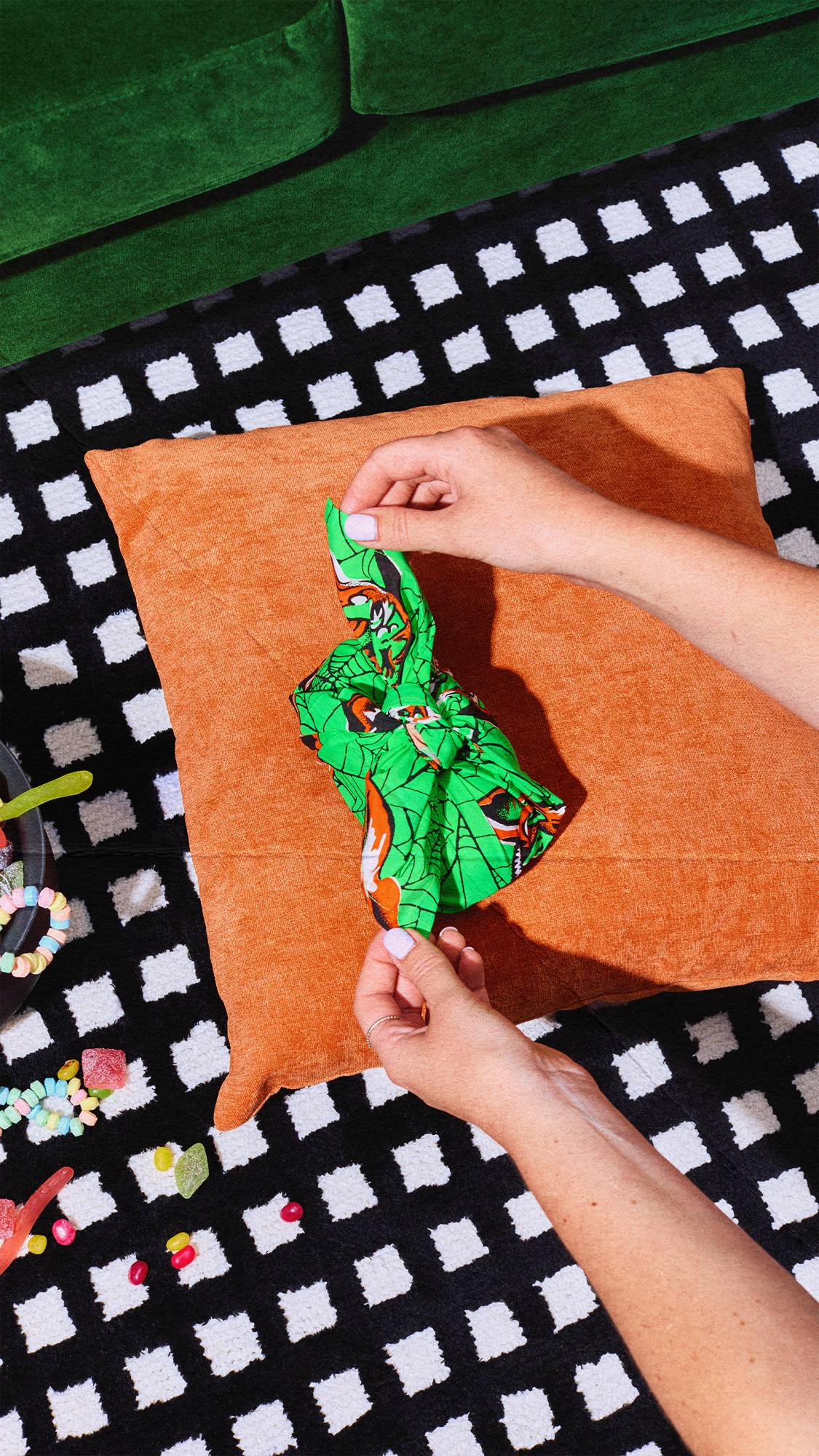 A person's hands untie a small gift wrapped in a vibrant green Halloween knot-wrap fabric. The gift rests on an orange cushion on a black-and-white chequered rug. Colourful sweets are scattered nearby, with a green couch at the top.