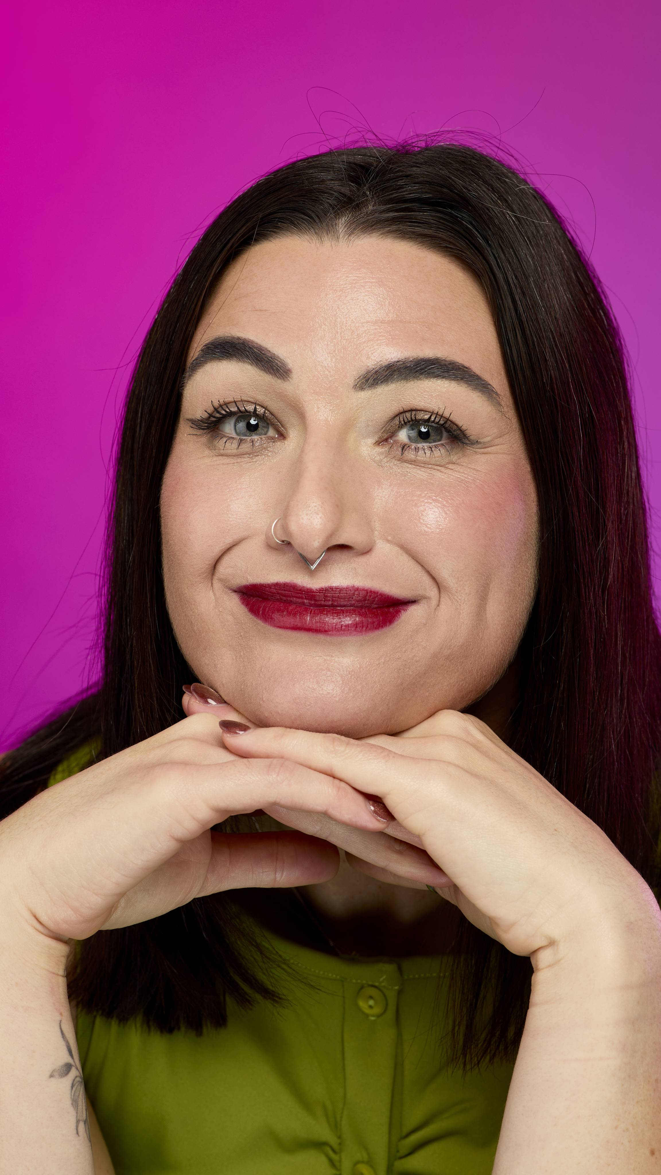 A person with dark hair, wearing a green top and a septum ring, smiles with deep crimson red lips (consistent with the "Crimson Reds" Lipstrick Square). They rest their chin on their hands against a bright magenta background.