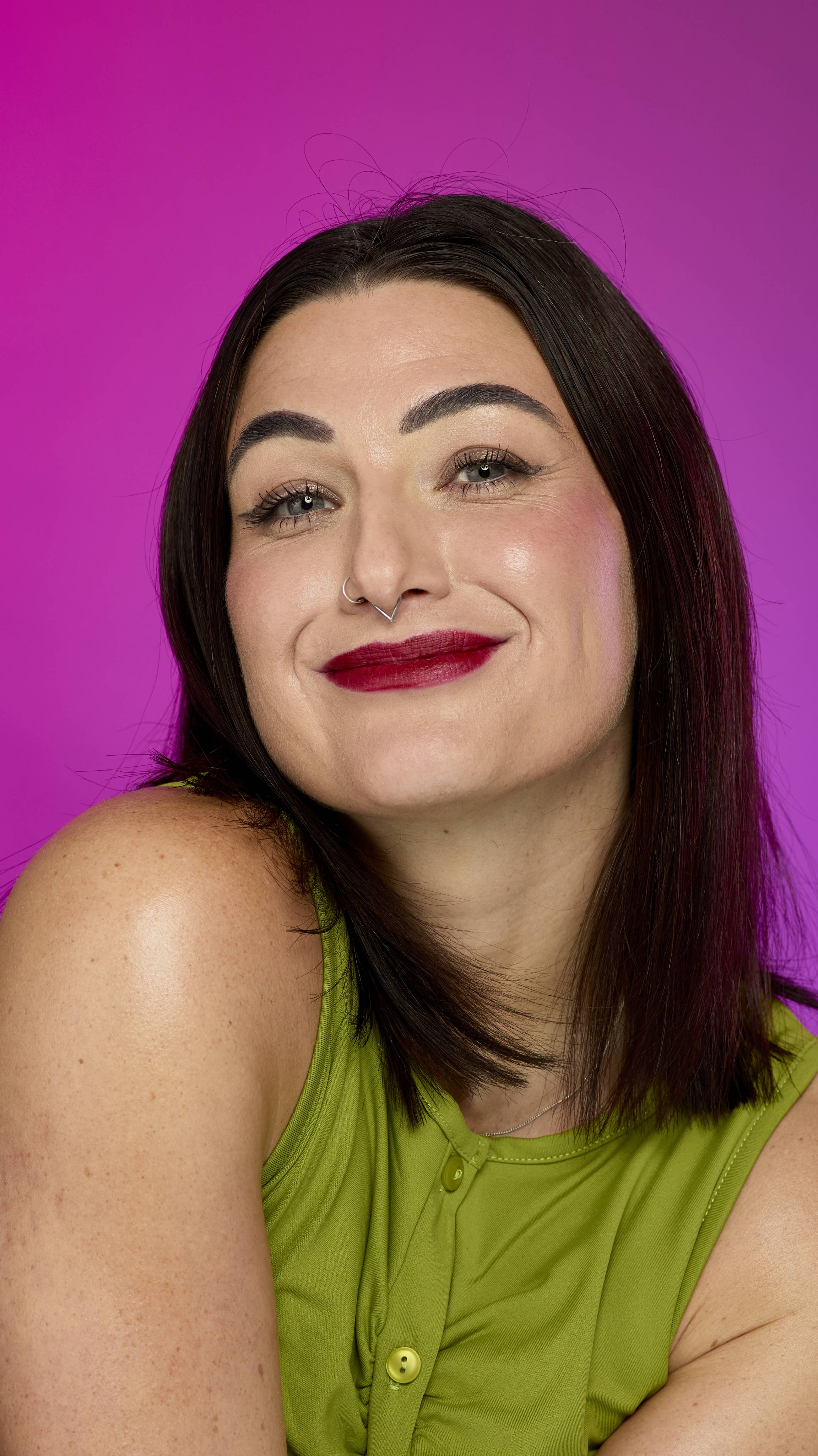 A person with dark hair, wearing a chartreuse green top and a septum ring, smiles directly at the camera. They wear a deep crimson red lip color consistent with the "Crimson Reds" Lipstrick Square, against a bright magenta background.