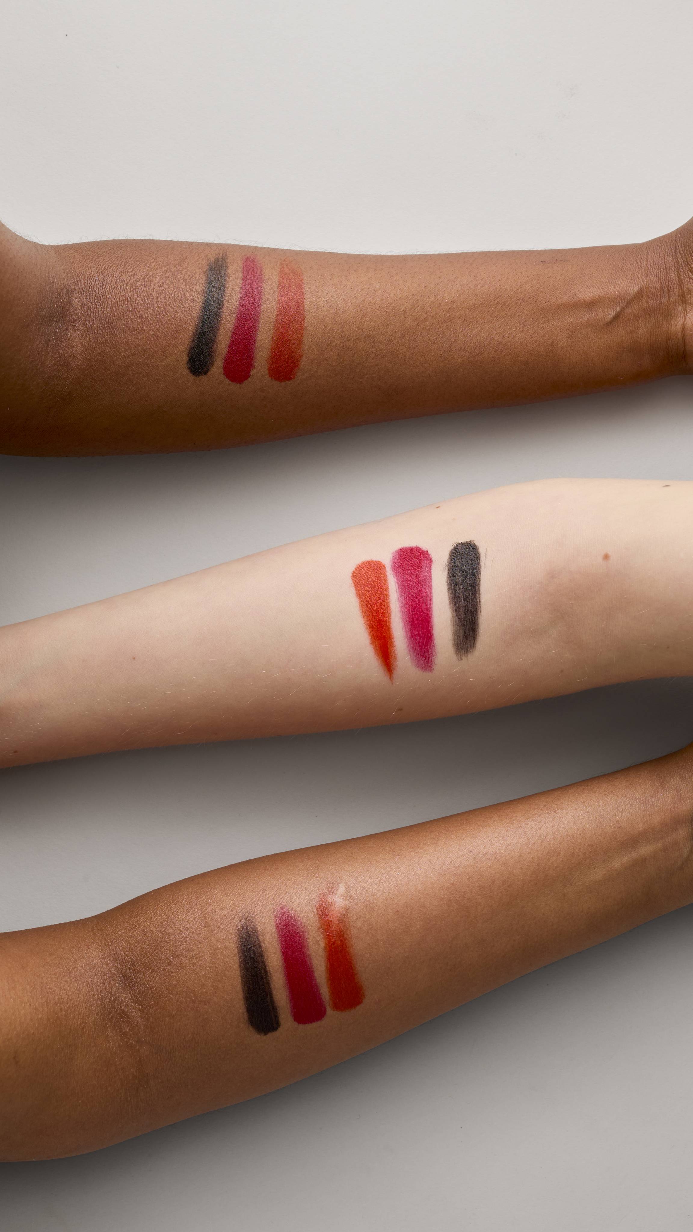 Three forearms with various complexions are shown on a light grey background. Each has three vertical swatches of Lush Lip square swatches: black, deep crimson red, and a vibrant orange/magenta/red-orange.