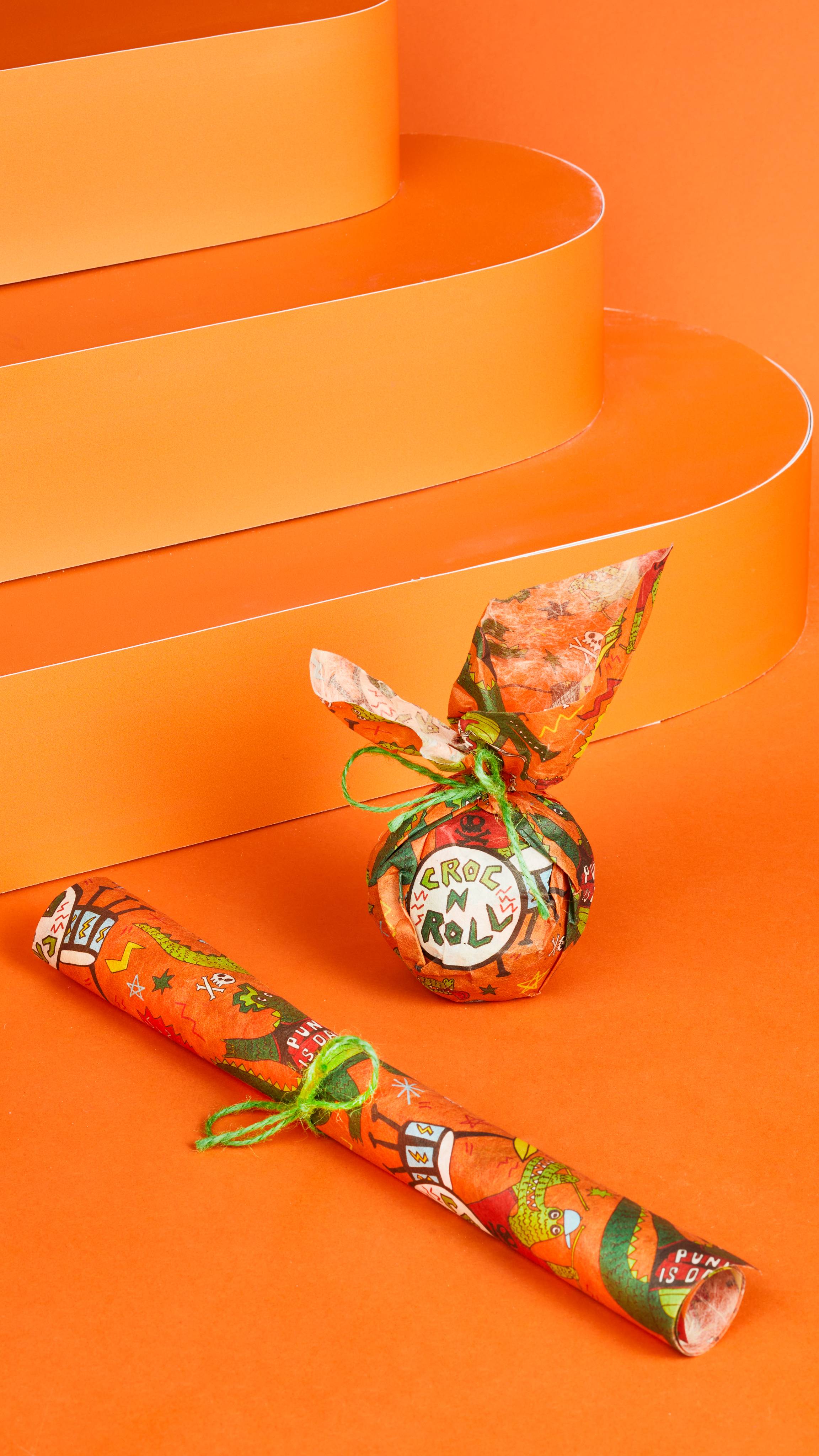 Croc And Roll Lokta Wrap displayed two ways, rolled up with string or wrapped around a gift, on a bright orange background.
