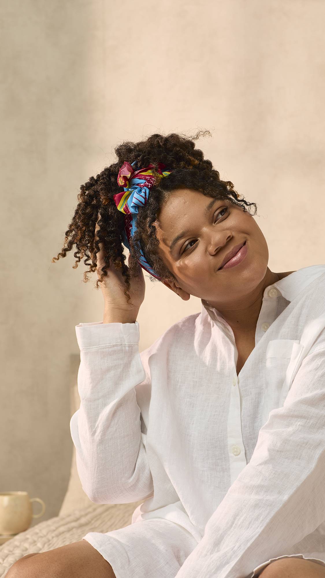 The model is sat on the bed in an oversized white shirt. They have fashioned the Curly Coily gift knot wrap into a trendy head bandana for their thick, curly hair. 