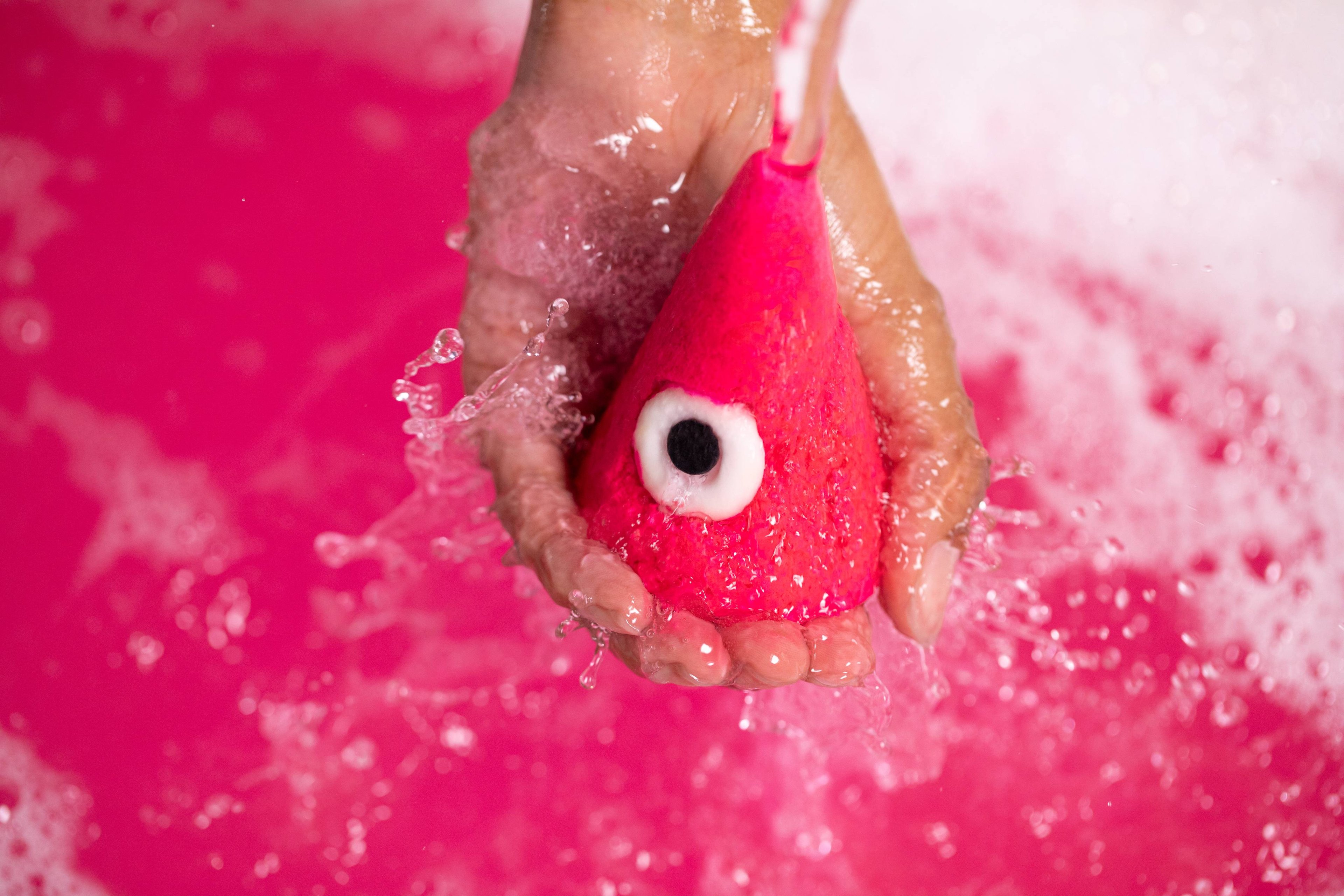 A hand holds the bubble bar under running water, lots of bubbles foam below on top of pink bath water.