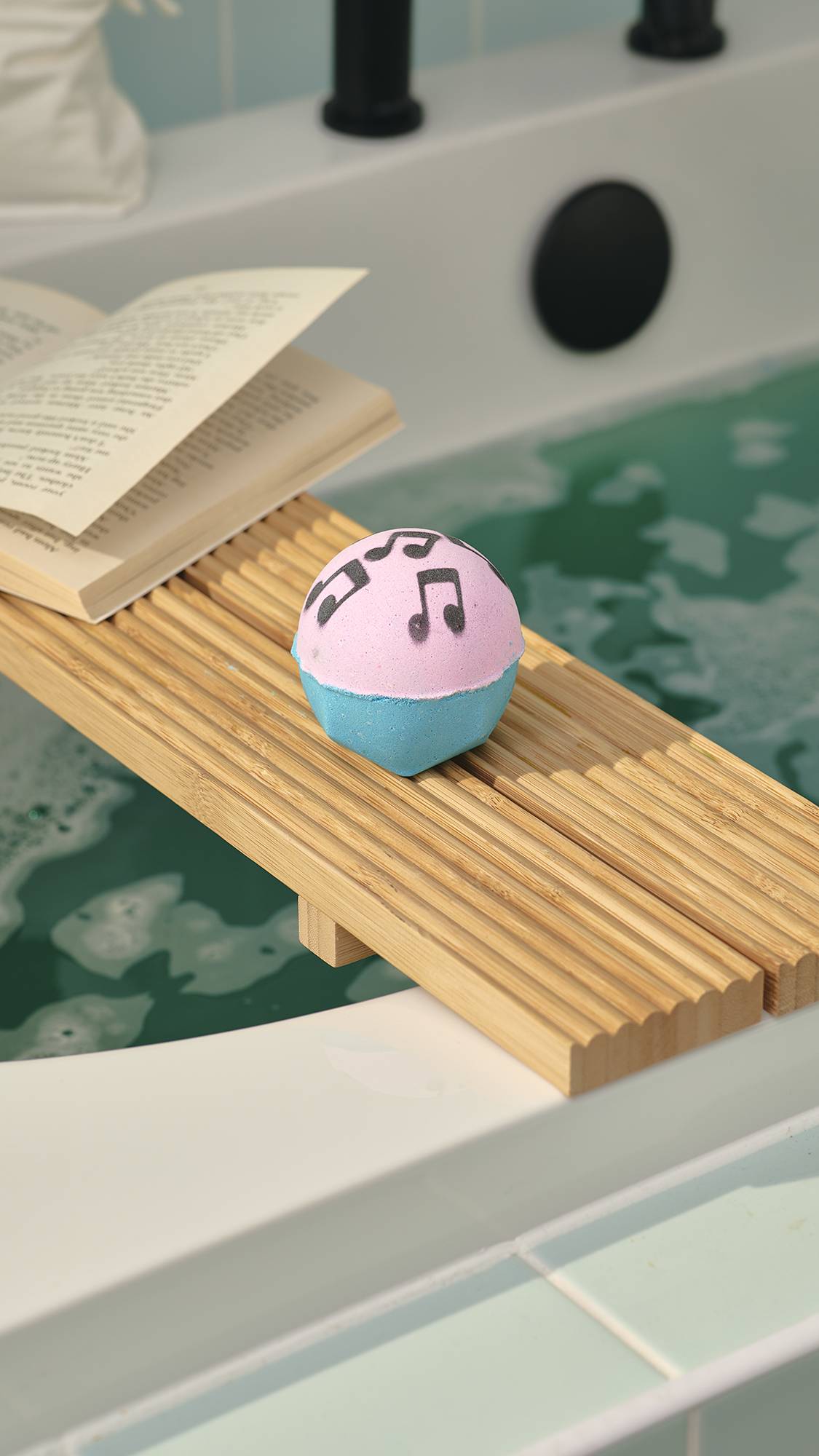 A pink and blue "Dad Dancing" bath bomb with black musical note stencils sits on a wooden bath tray over a tub of green water. A book rests next to it. In the background, a white bath with a black tap is set against pale green wall tiles.