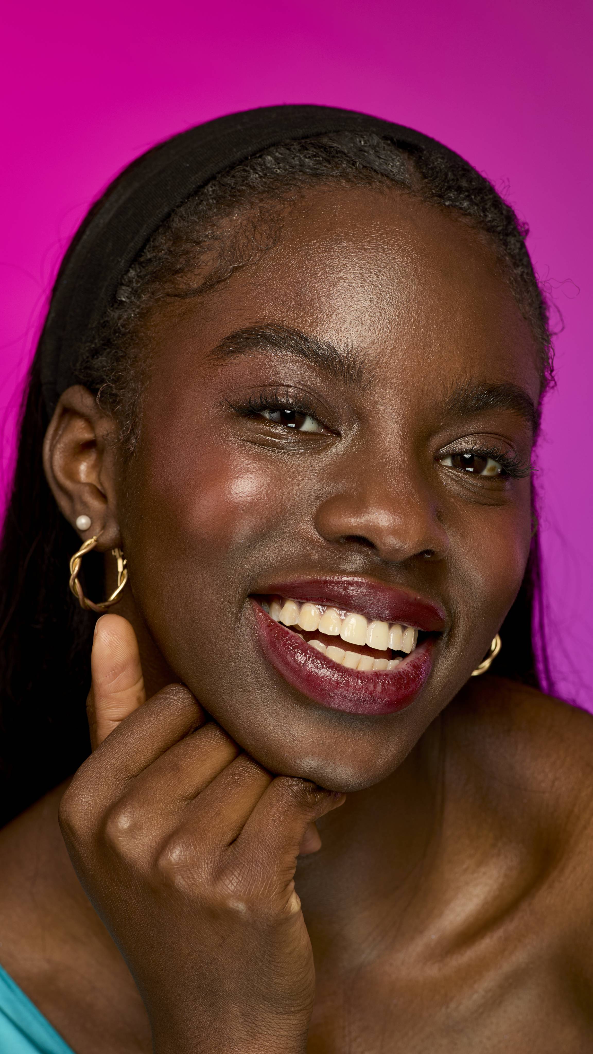 The image shows a model with long, dark hair on a vibrant magenta background, from the neck up. They are smiling widely while wearing the rich, warm, mahogany-brown Dahlia lipstick.