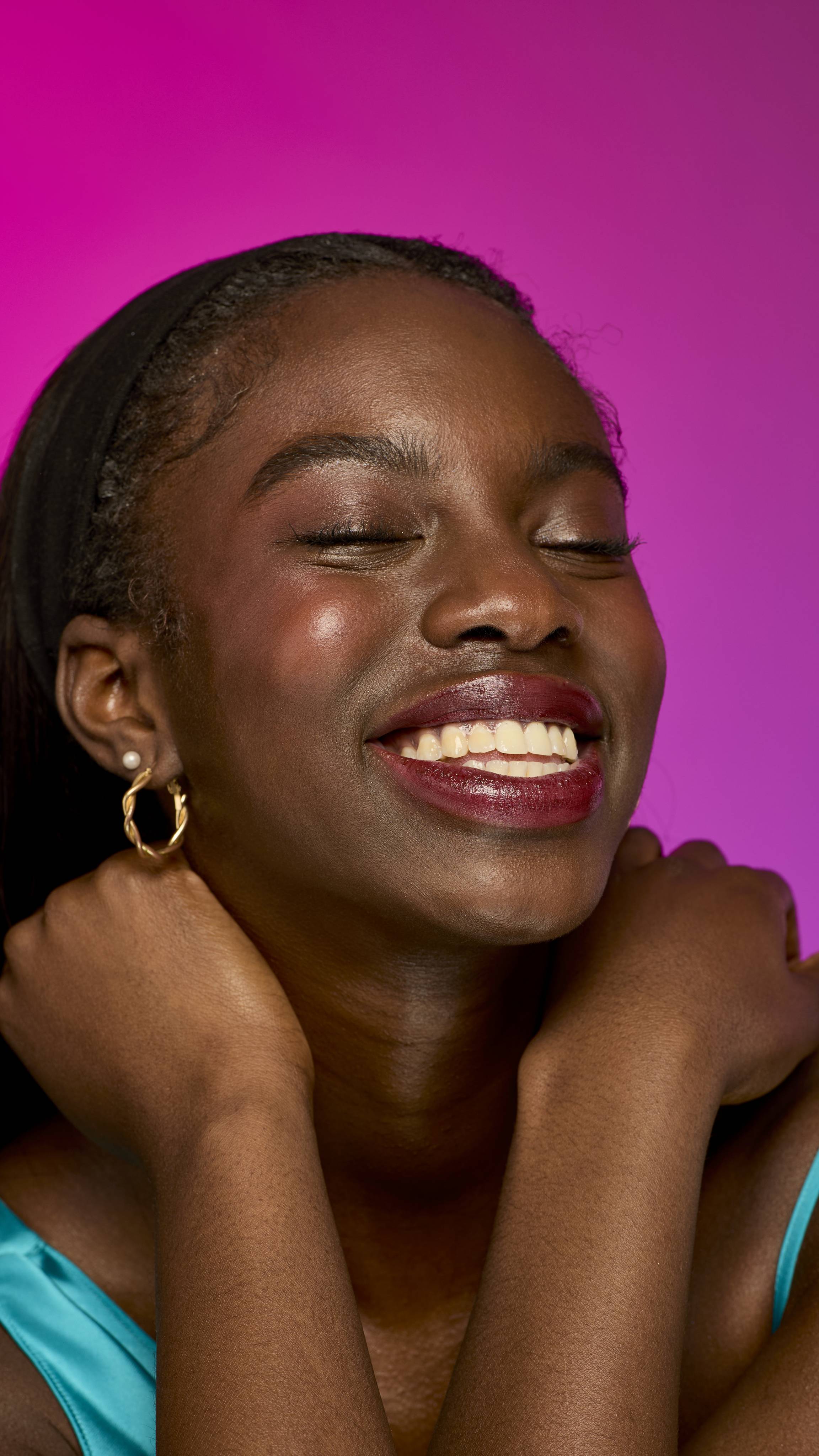 The image shows a model with long, dark hair on a vibrant magenta background, from the neck up. They are smiling widely while wearing the rich, warm, mahogany-brown Dahlia lipstick.