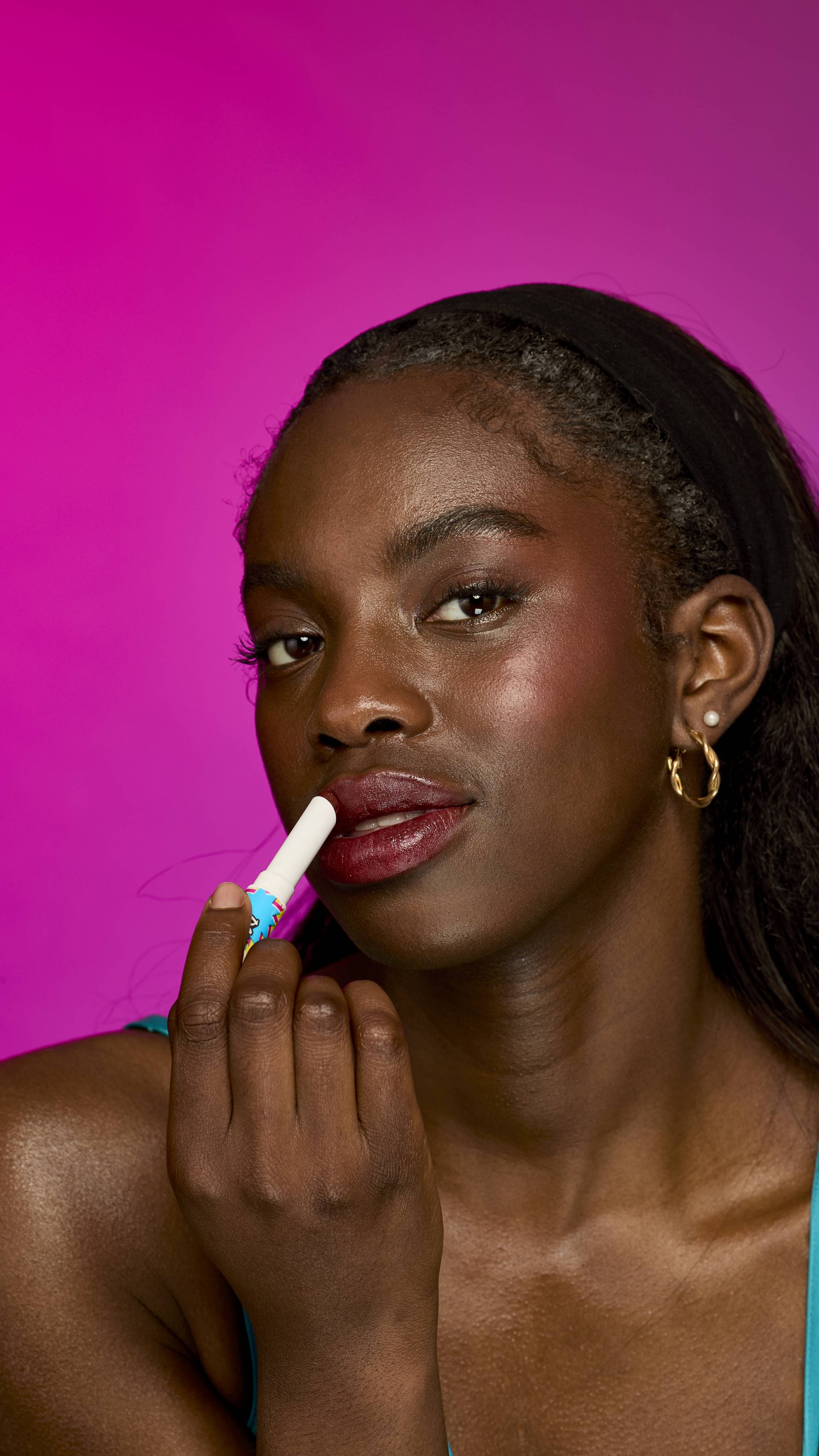The image shows a model with long, dark hair on a vibrant magenta background, from the neck up. They are applying the rich, warm, mahogany-brown Dahlia twist-up lipstick.