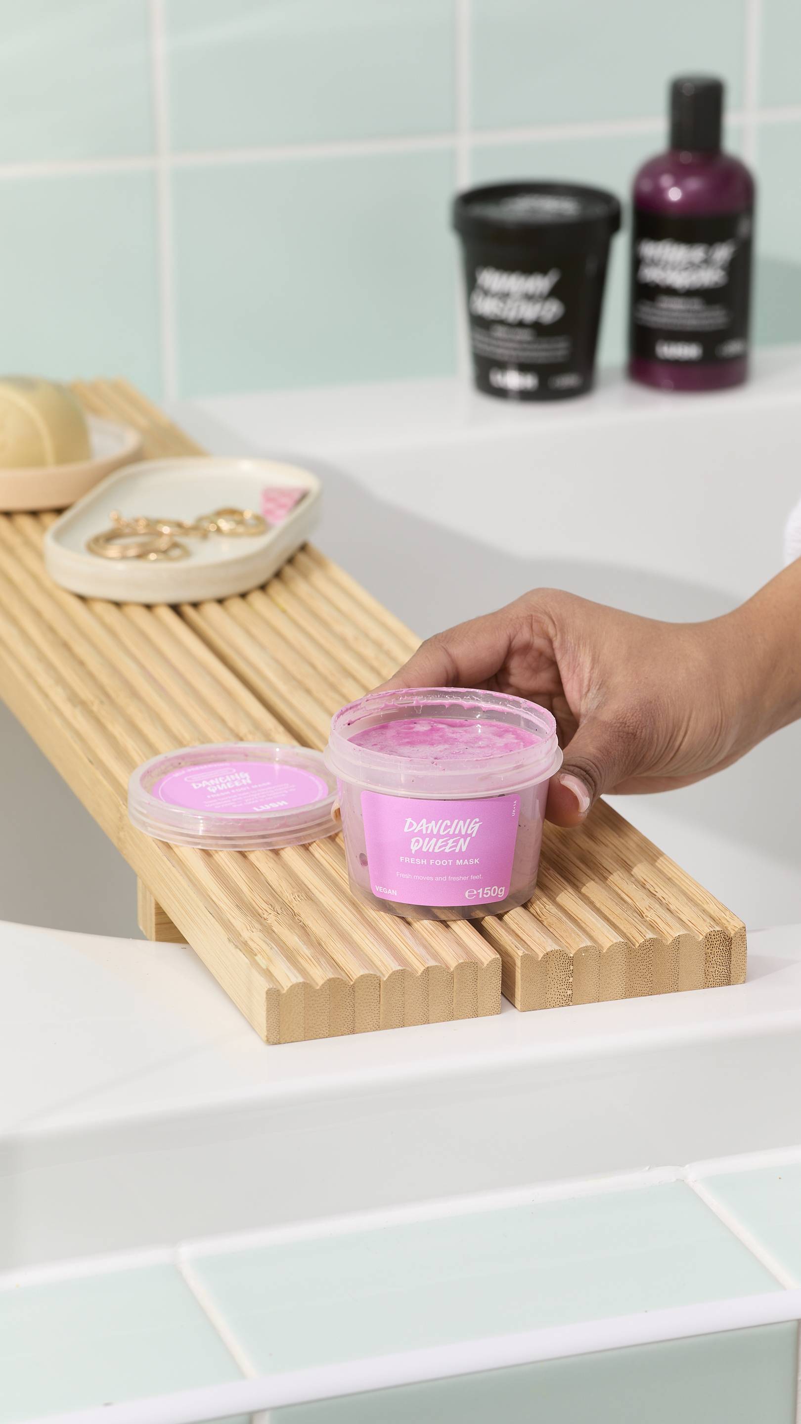 A hand reaches for an open tub of "Dancing Queen" pink foot mask sitting on a wooden bath tray. The background features a white bathtub, light green wall tiles, and other LUSH self-care products.