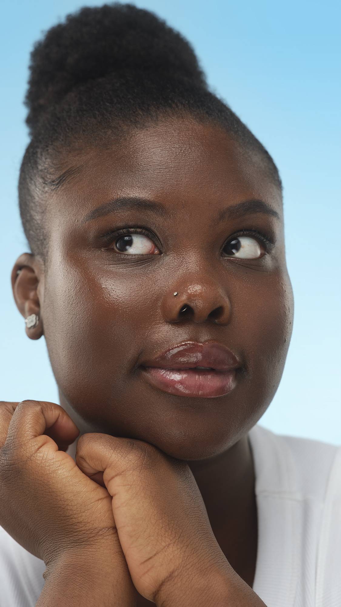 A close-up portrait of a person with deep skin, dark hair in a bun, and a nose stud, resting their chin on clasped hands. They look up and right, with a luminous finish from the Dark Cool Skinwear, against a bright blue background.