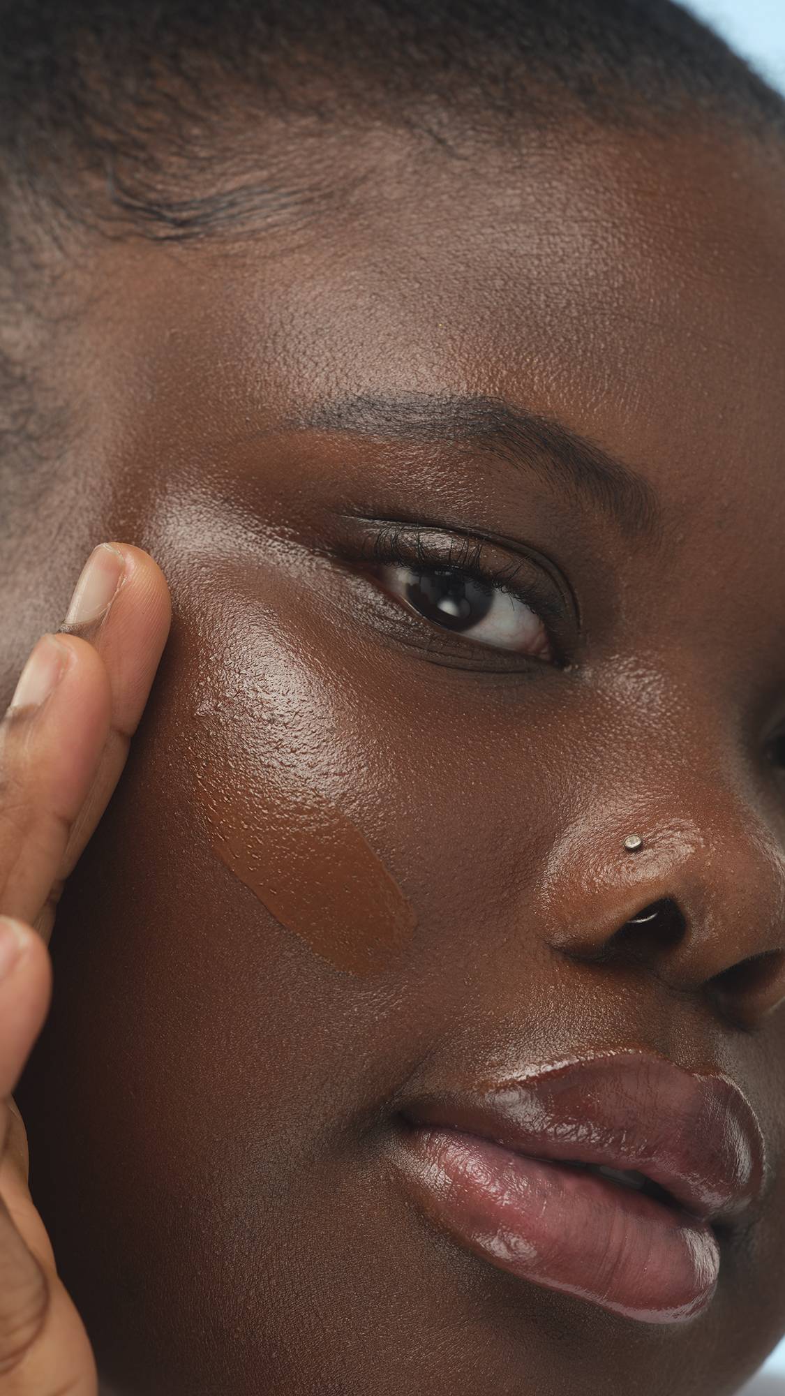 A close-up vertical shot of a person with deep skin, looking at the camera while applying a thick swatch of Dark Cool Skinwear to their cheek with a finger. The skin has a luminous finish and they wear a nose stud.