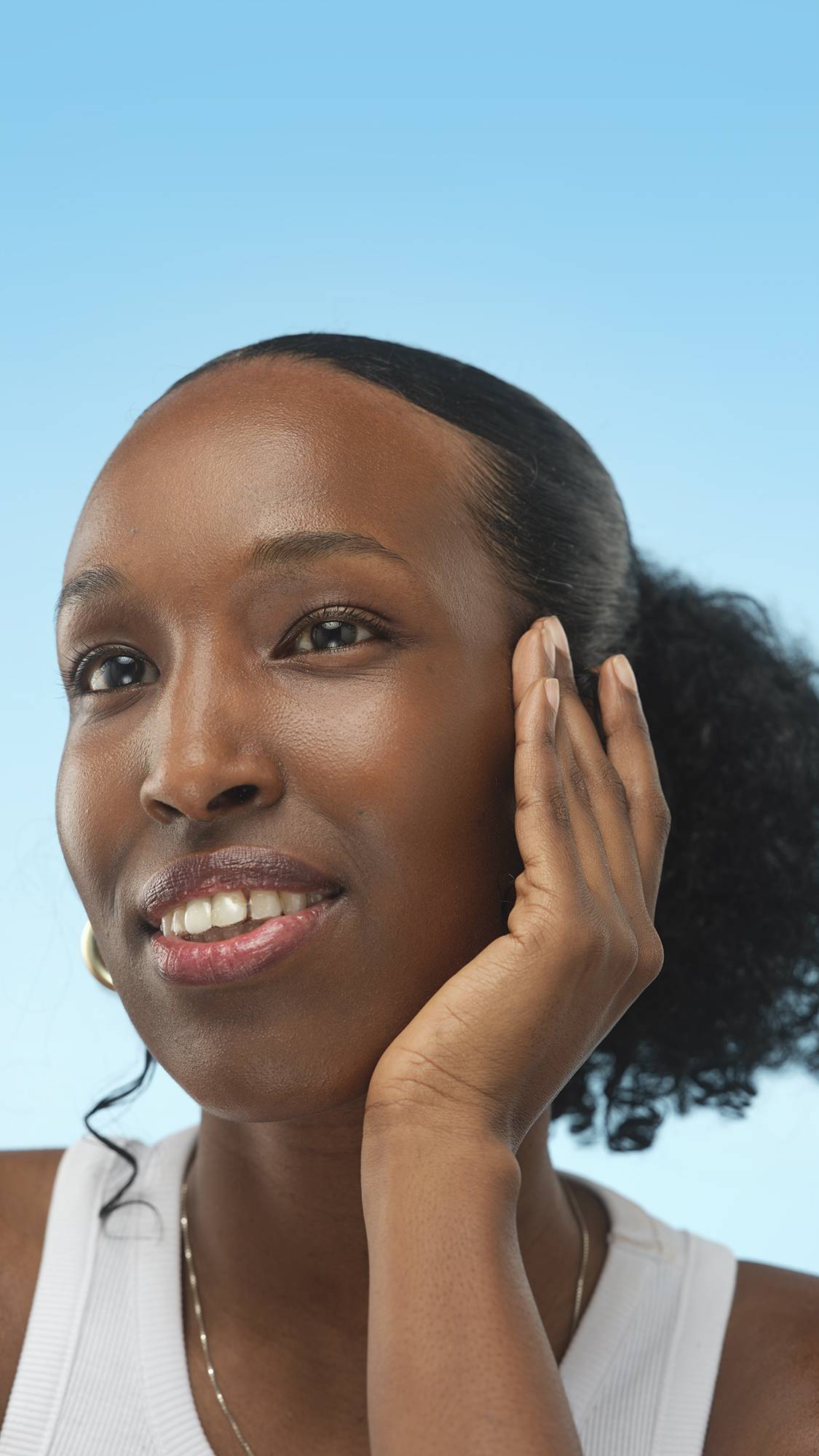 A person with dark, warm skin smiles and rests a hand on their cheek against a light blue background. The person is wearing a white top and a delicate silver necklace.