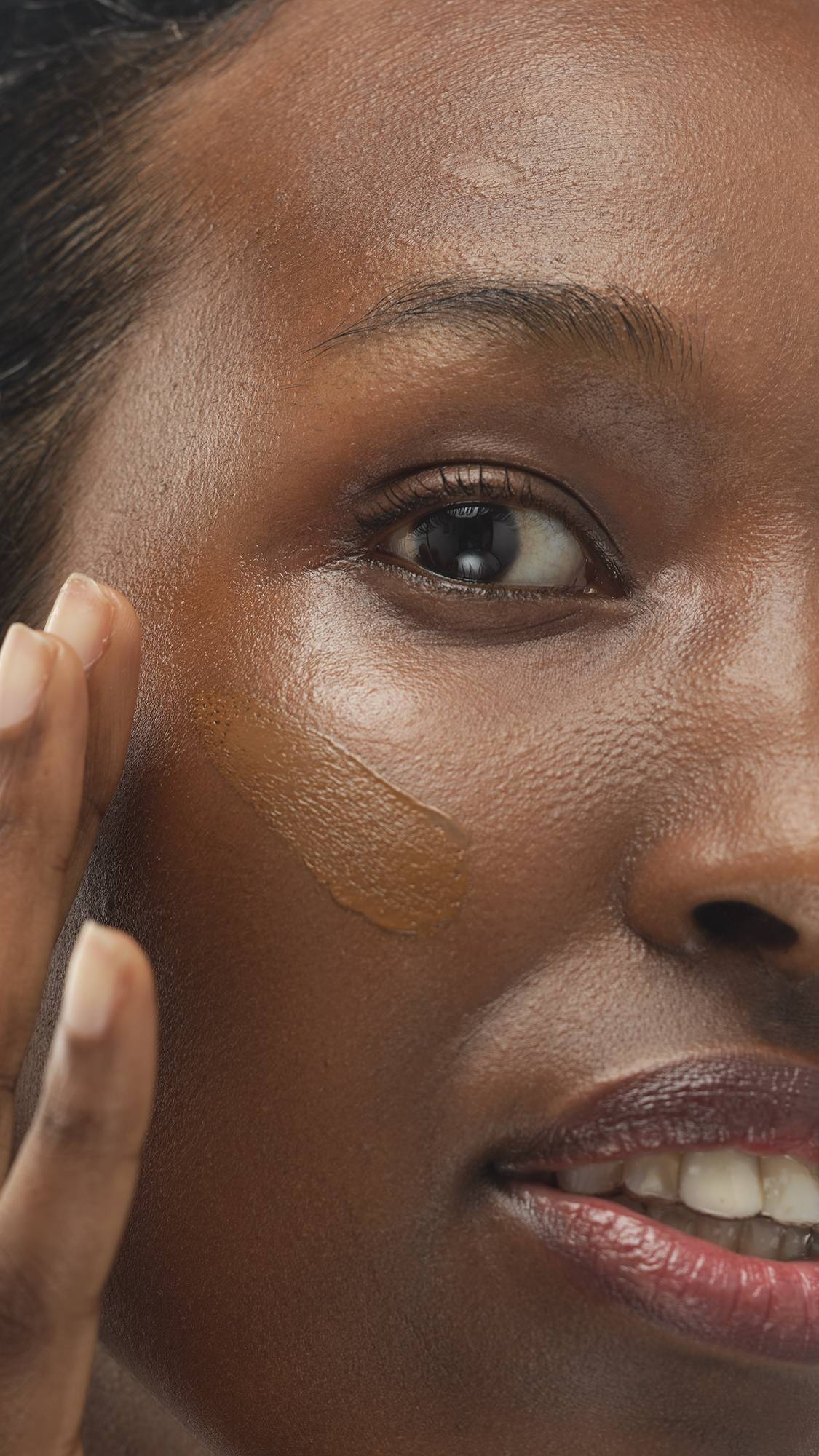 Close-up on a person's face with dark, warm skin as they use their finger to apply a tan foundation or skin tint in a thick vertical swatch across their cheek.