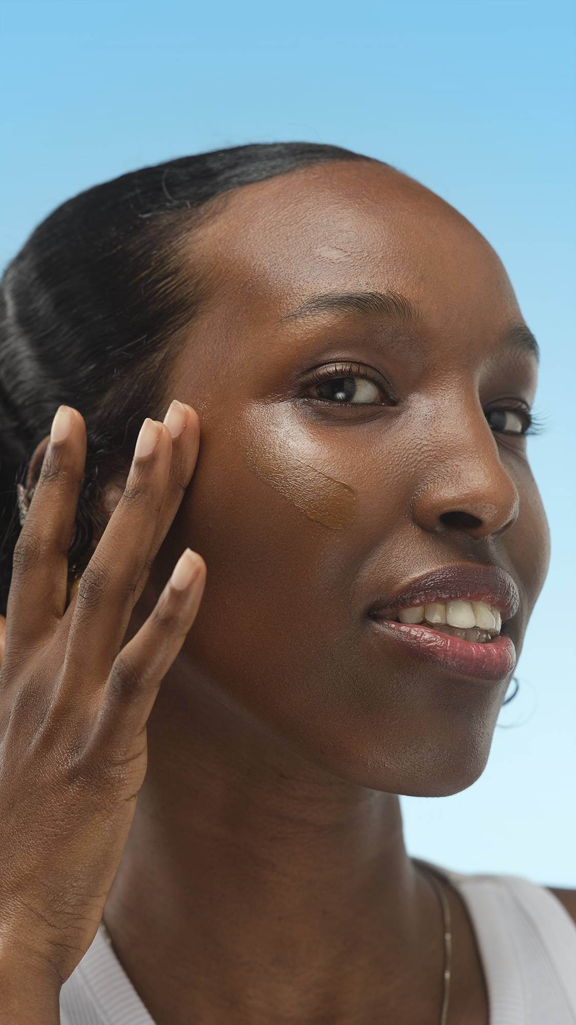 A smiling person applies a dark brown foundation or skin tint with their finger, swiping it across their cheek. The person has dewy skin and is posing against a light blue background.