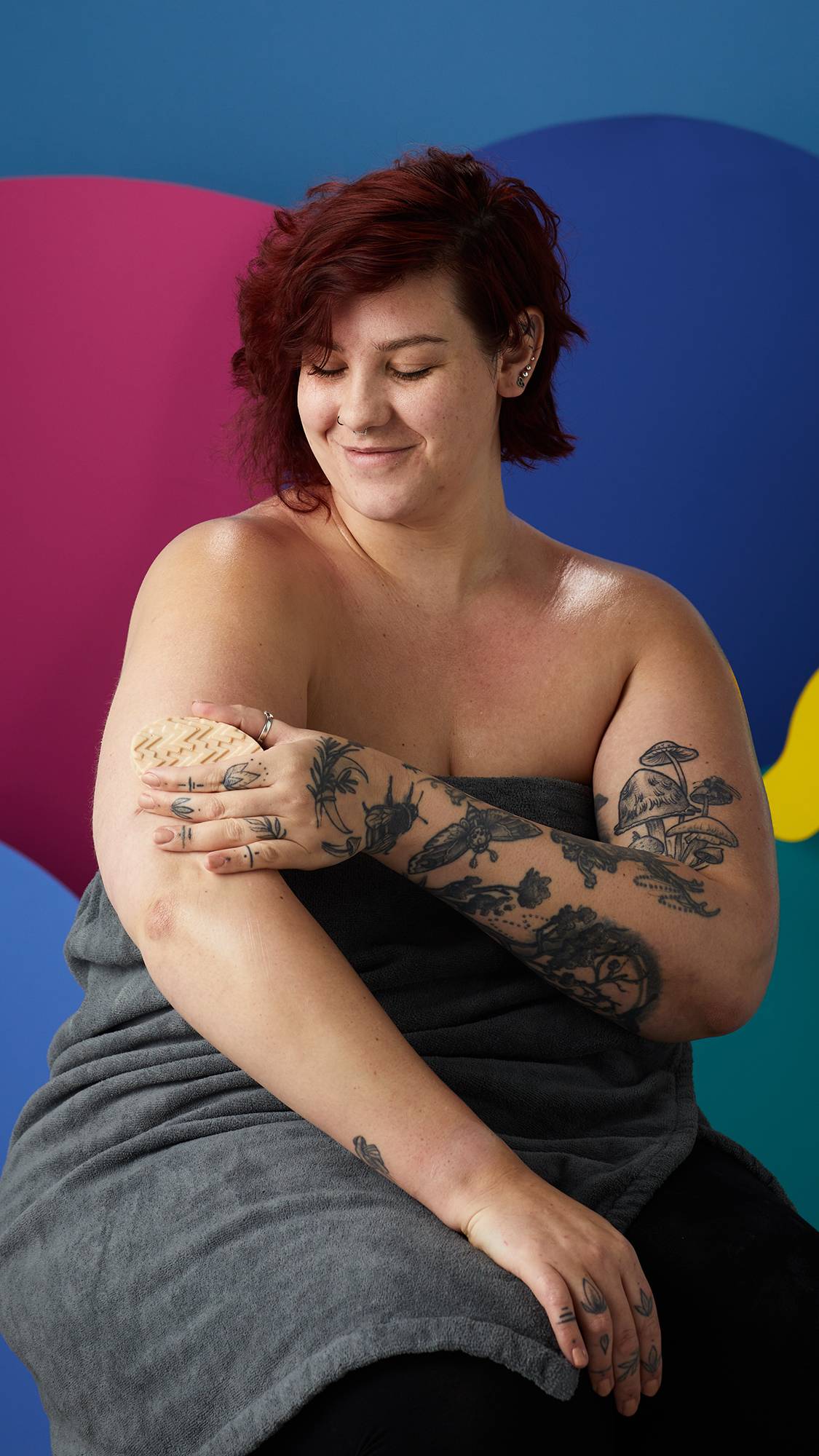 A model with red hair and tattoos sits wrapped in a grey towel, smiling. They are using a "Deep Sleep" magnesium massage bar on their upper arm against a vibrant, colourful backdrop. 