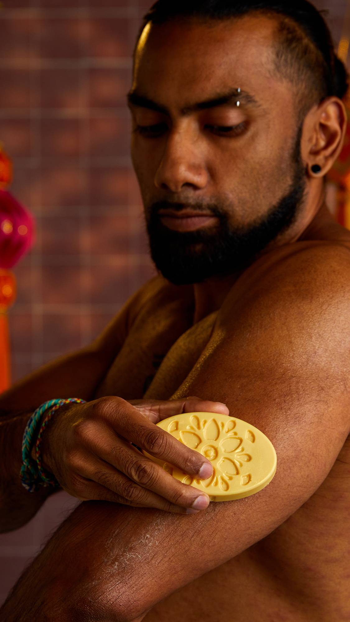 A person with a beard and tattoos holds a solid moisturiser in his hand, rubbing it on his bicep. The solid moisturiser is a light yellow oval with a floral design pressed into it.