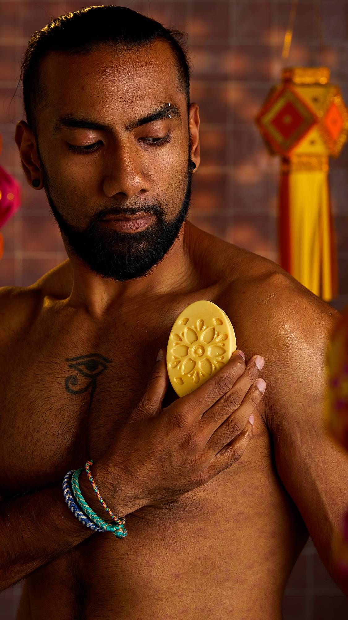A person with a beard and tattoos holds a solid moisturiser in his hand, rubbing it on his bicep. The solid moisturiser is a light yellow oval with a floral design pressed into it.