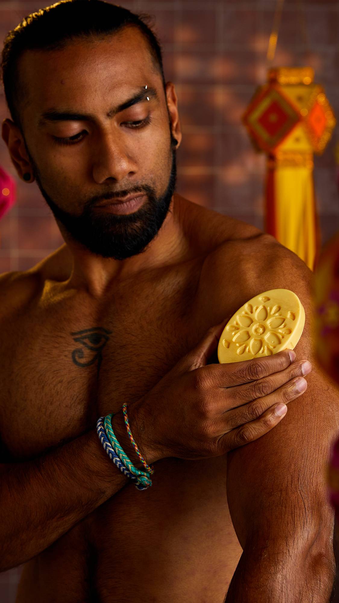 A person with a beard and tattoos holds a solid moisturiser in his hand, rubbing it on his bicep. The solid moisturiser is a light yellow oval with a floral design pressed into it.