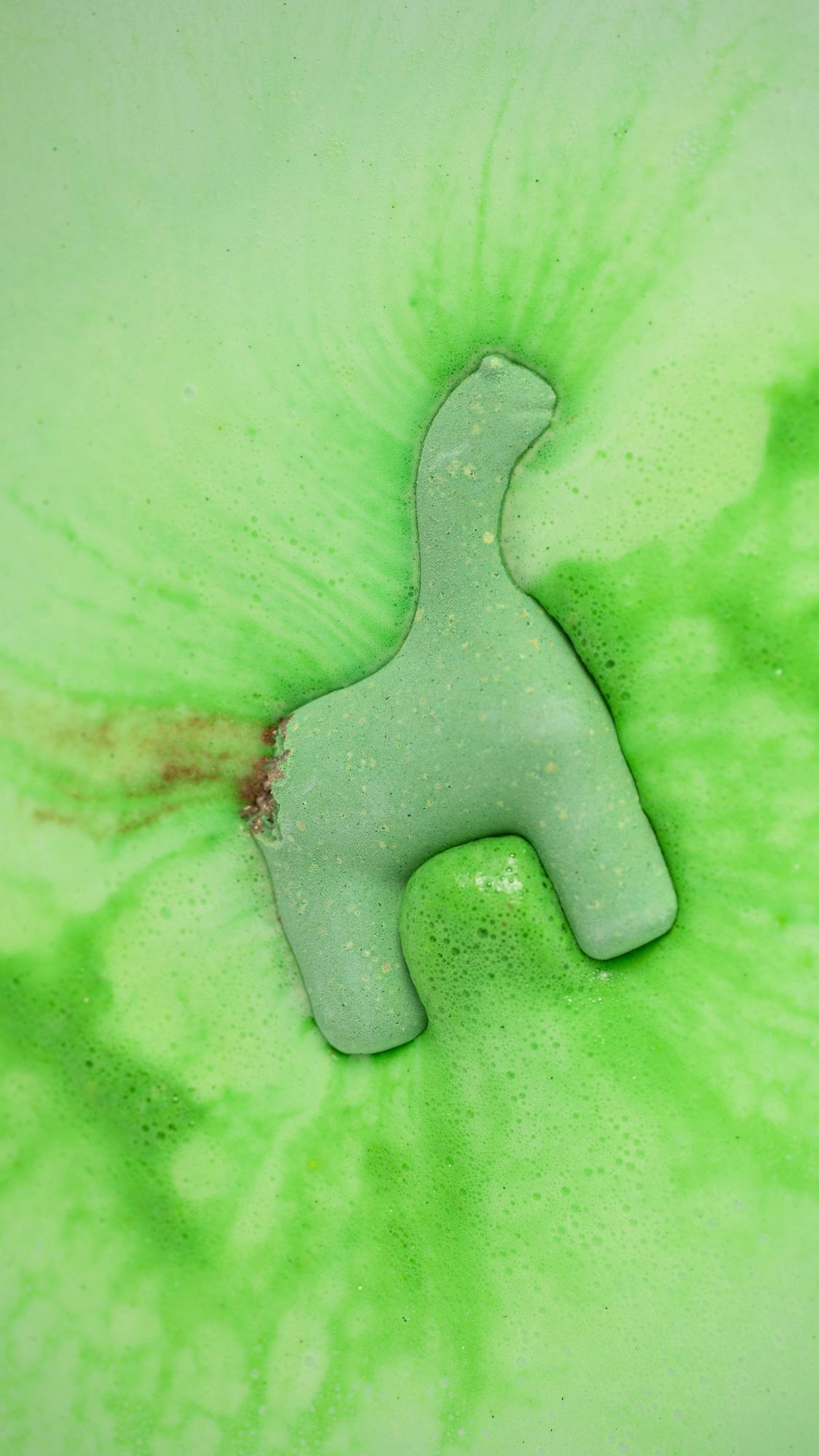 The green Dinosaur in a Crisis bath bomb is sitting dissolving in the water surrounded by thick, green foam and a hint of shimmering bronze. 