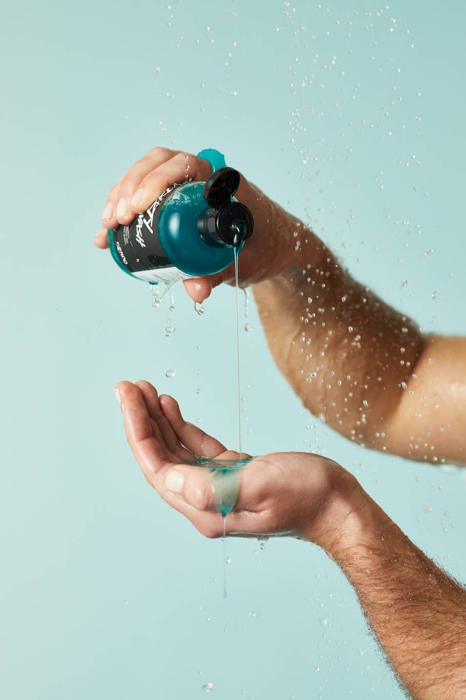 A close-up of the model's forearms on a pastel blue background as they gently squeeze the cool-blue Dirty Springwash shower gel into the palm of their hand under running shower water. 