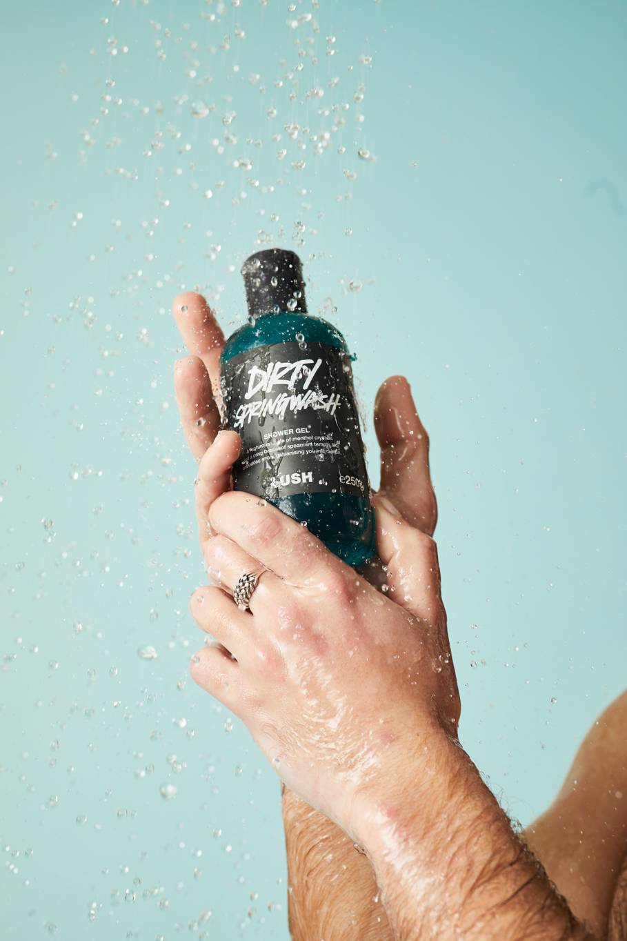 A close-up of hands holding the Dirty Springwash shower gel bottle under a shower stream on a light blue background.