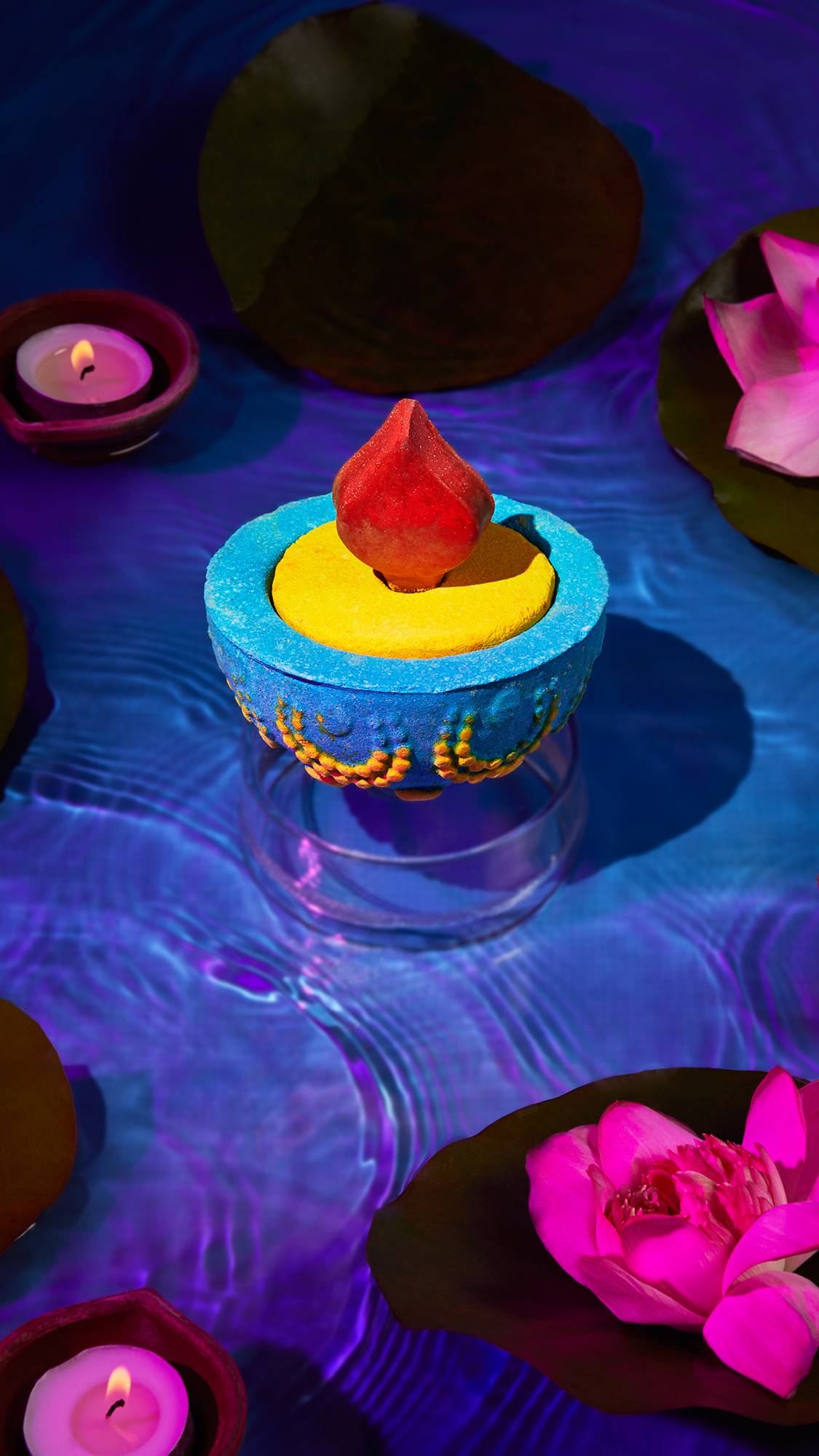 The image shows the Lush, Diwali-themed, Diya, 3-in-1 bath bomb hovering just above crystal clear water. There is a blue and purple hue as lotus flowers and small dishes with tealight candles float on the surface. 