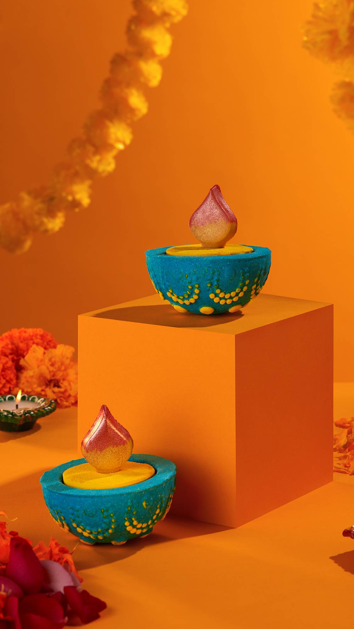 Image shows two Diya bath bombs placed around and on a pedestal. There is a vivid orange background with a marigold garland.